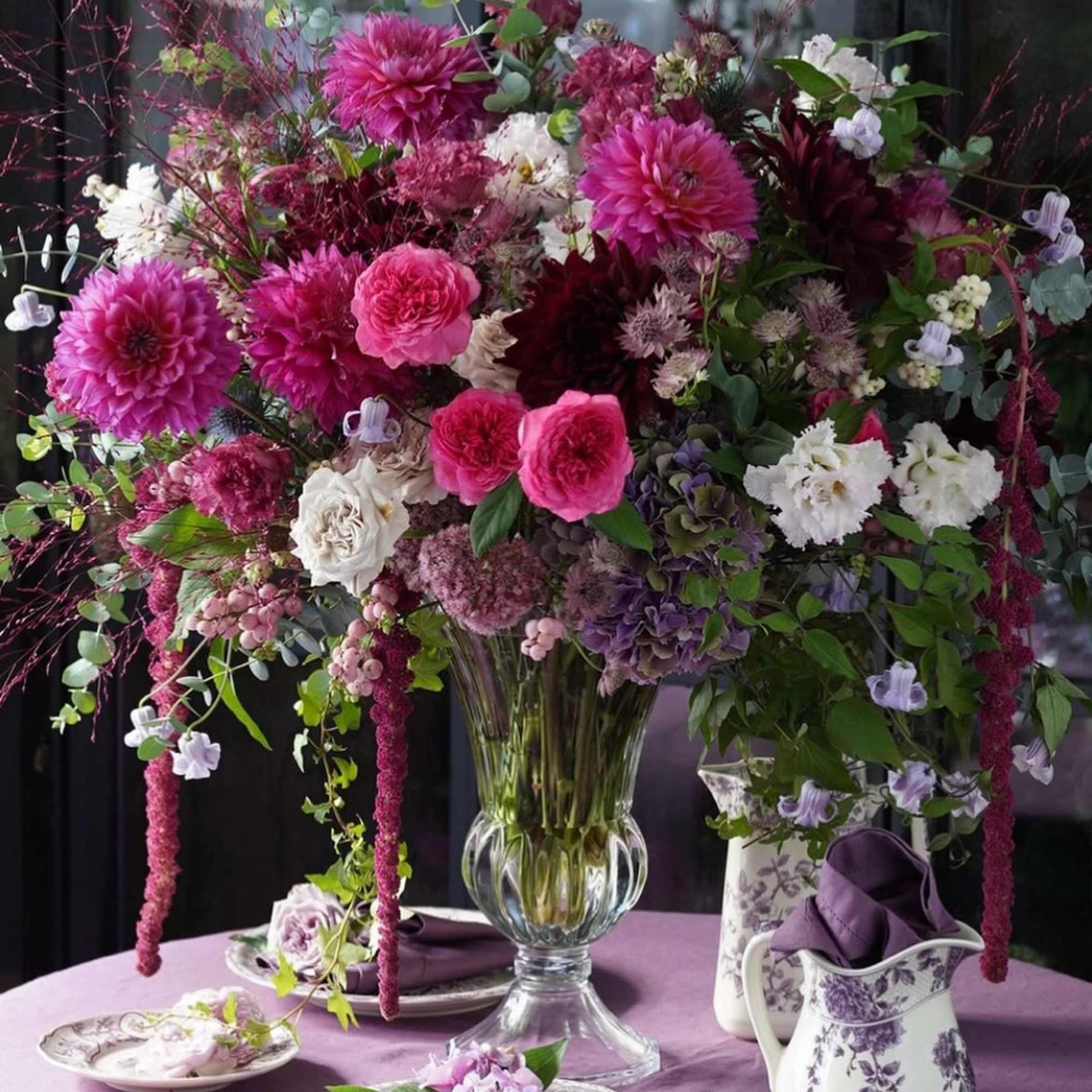 Large pink and white floral arrangement in a clear glass vase on a lavender tablecloth.
