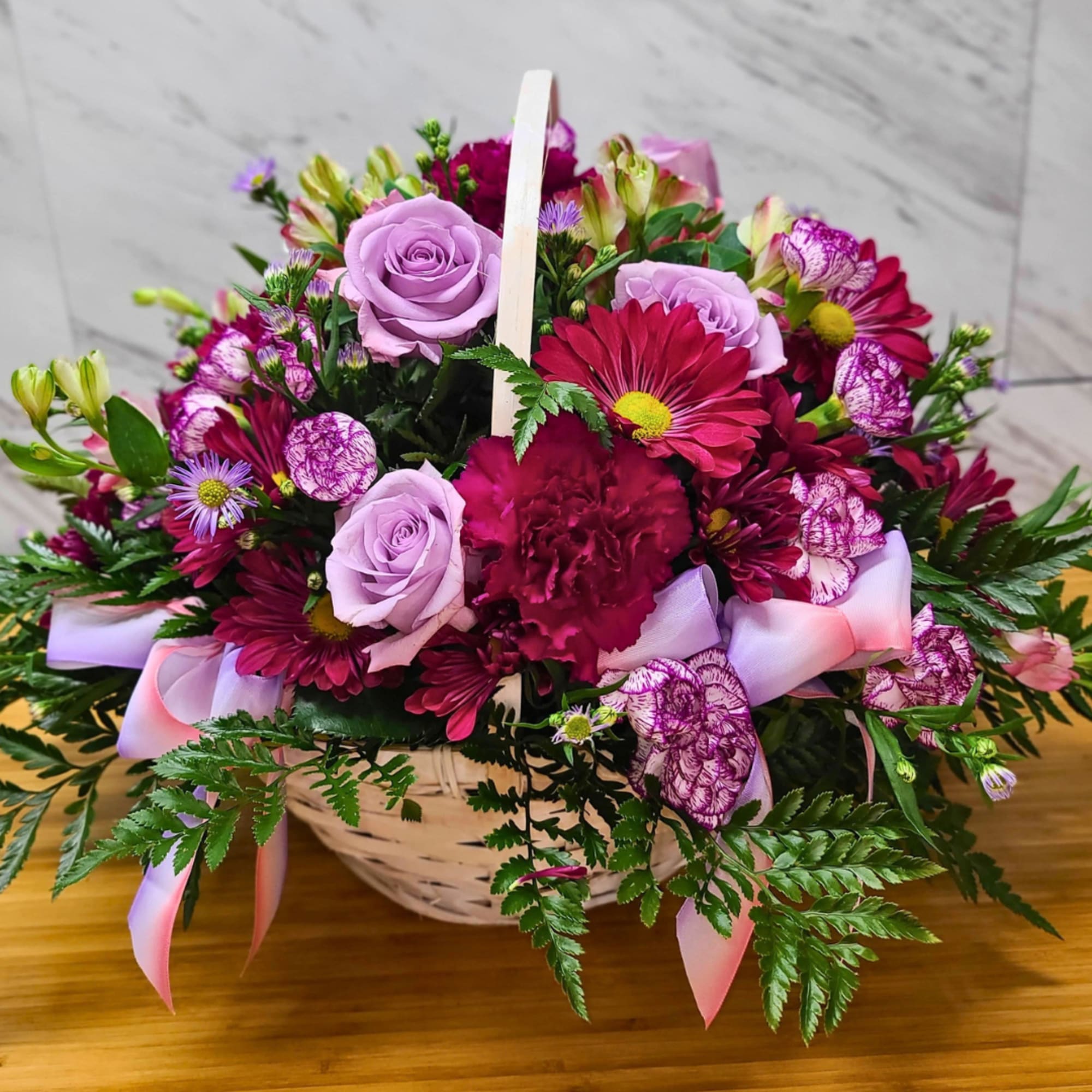 Basket arrangement of lavender roses, magenta daisies, and pink ribbon