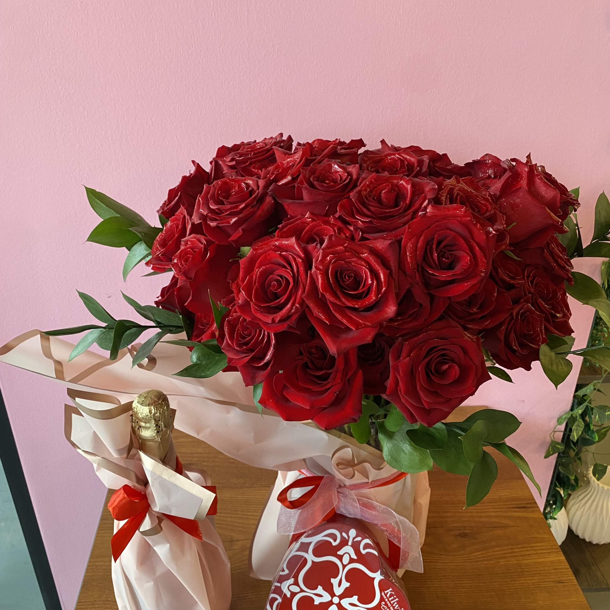 Bouquet of red roses with greenery, wrapped in pink paper beside a champagne bottle and heart box