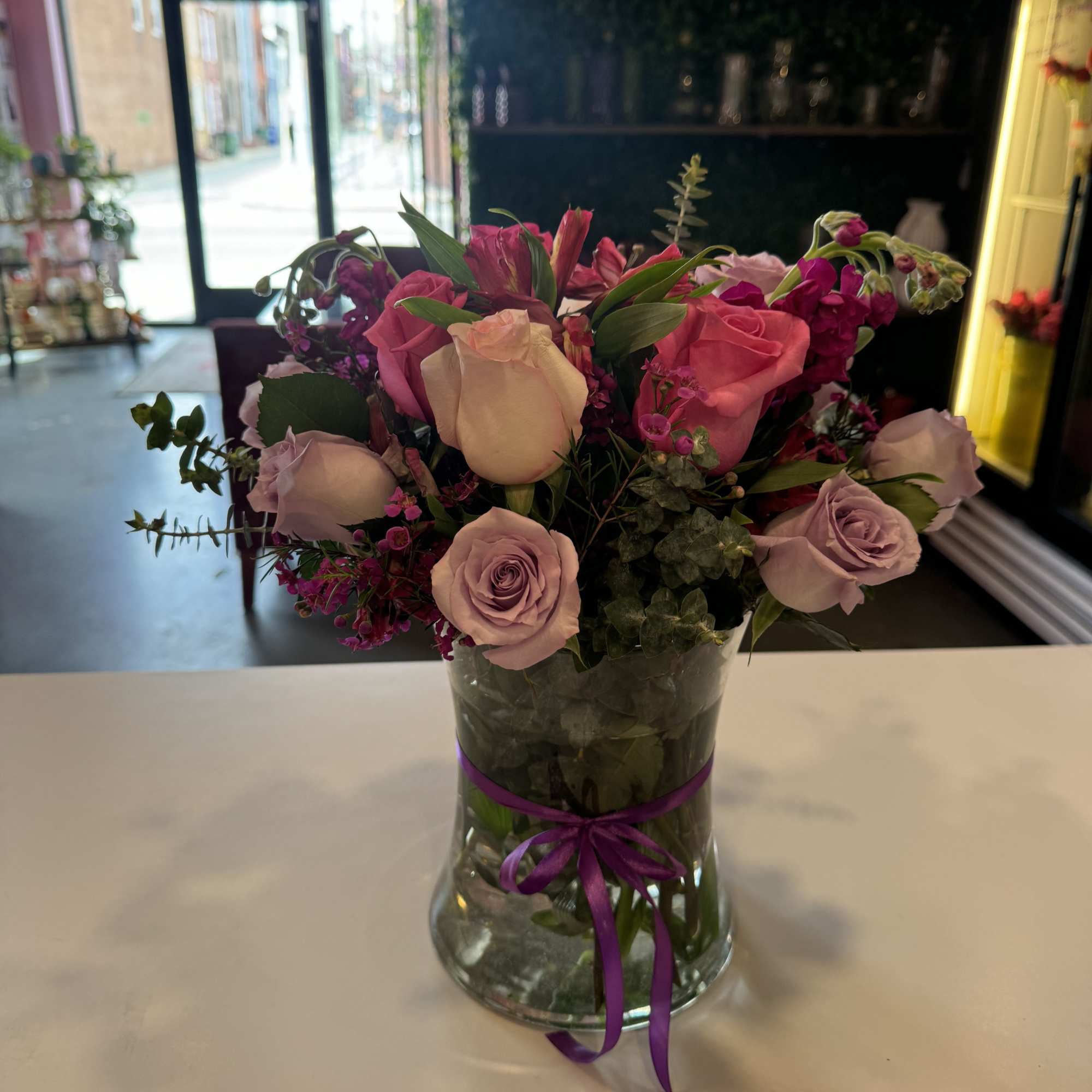 Pink and lavender roses arranged in a glass vase with a purple ribbon