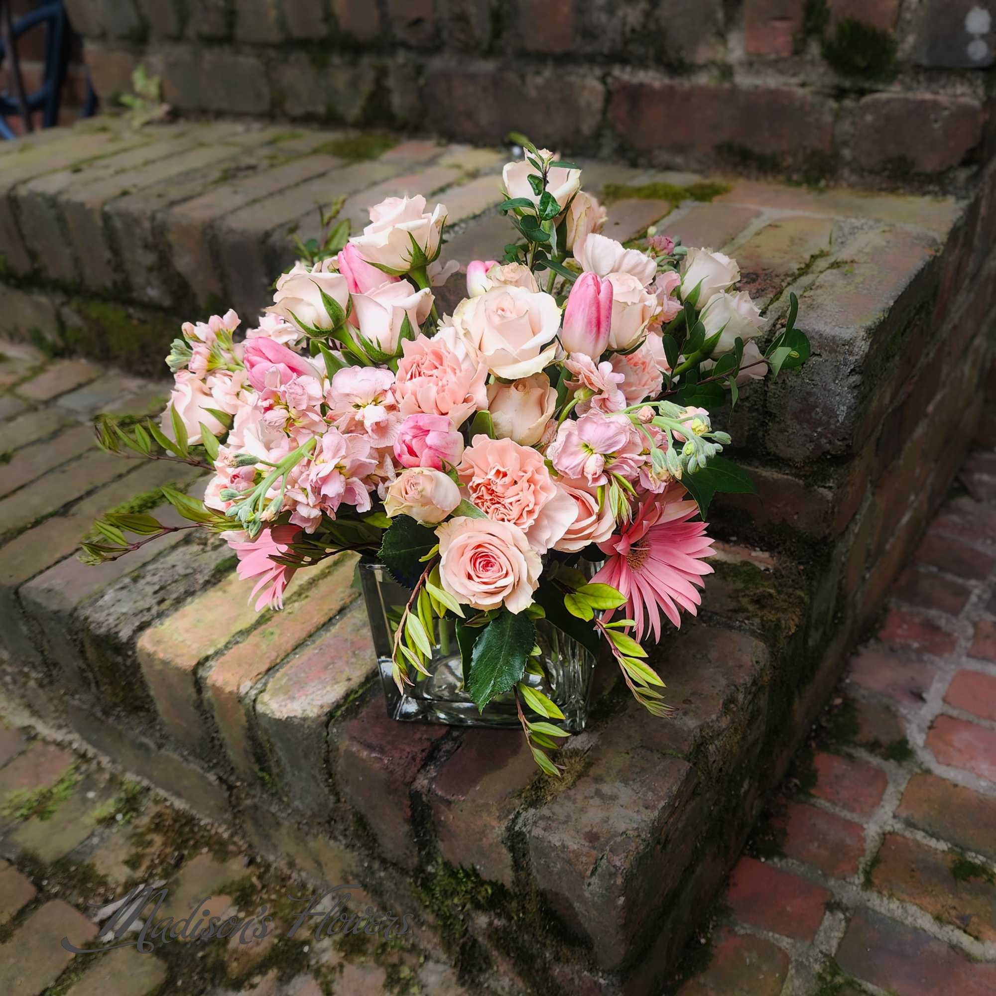 Low pink and peach flower arrangement with roses, carnations, and gerbera daisies in a glass cube vase.