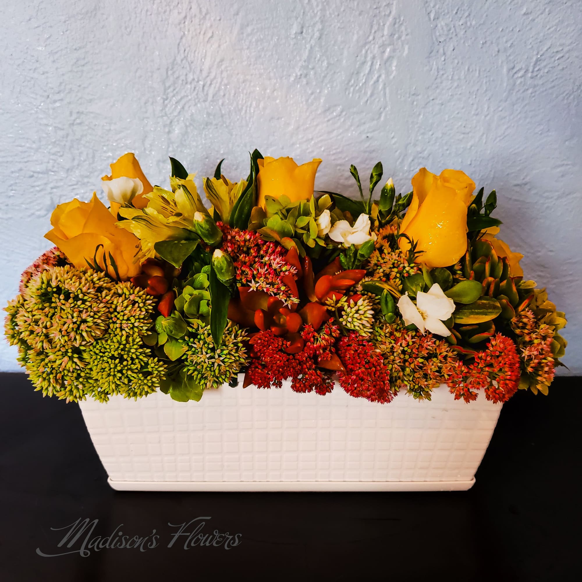 Yellow roses and mixed blooms arranged in a white rectangular planter