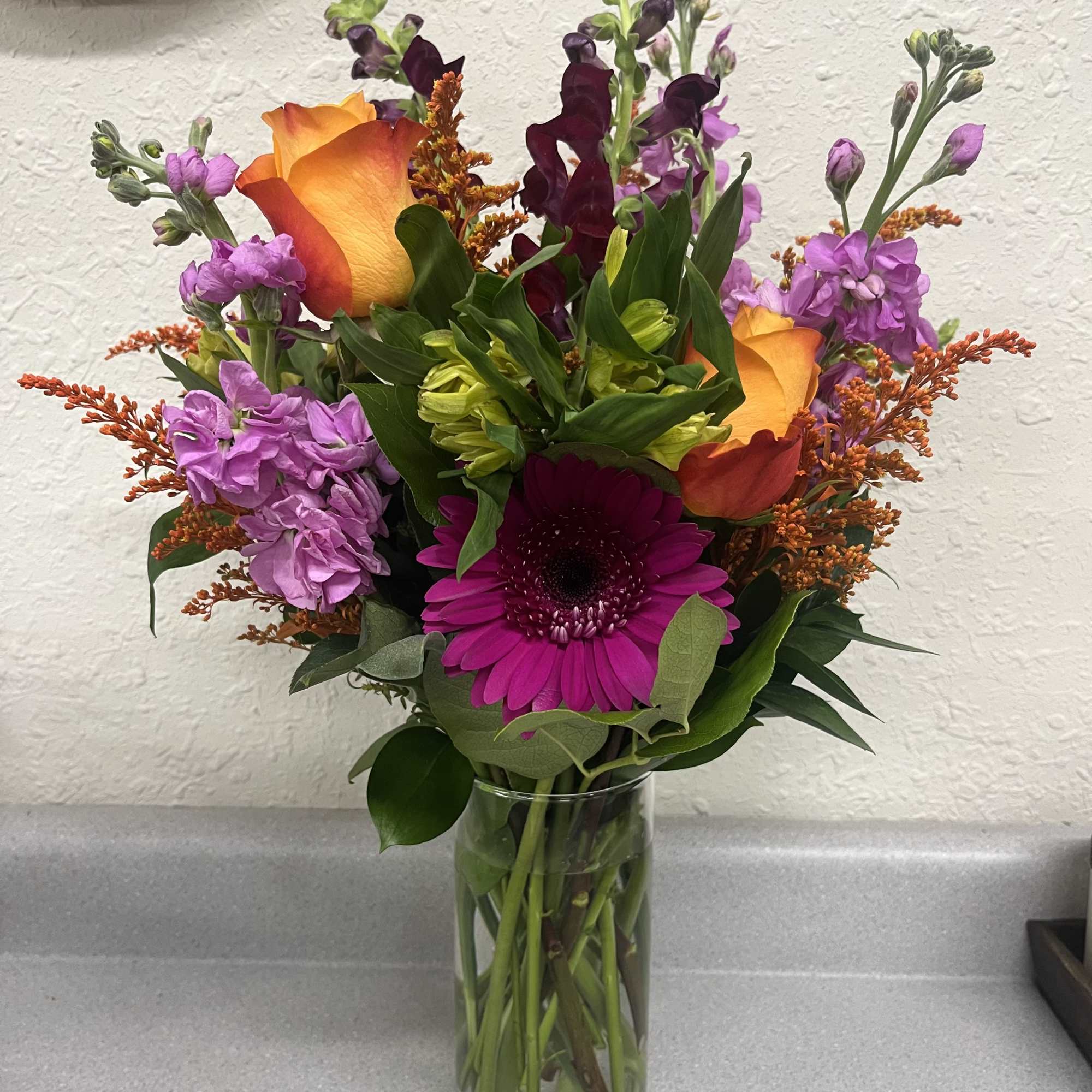 Mixed bouquet with orange roses, purple blooms, and a magenta gerbera in a glass vase