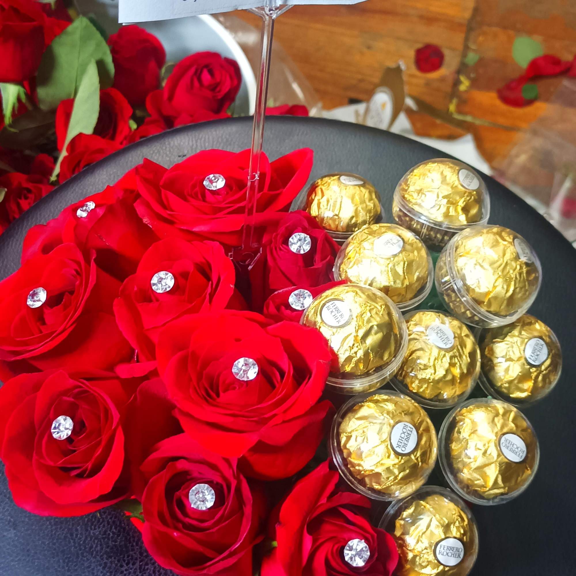 Red roses and gold-wrapped chocolates arranged on a black round box with a card.