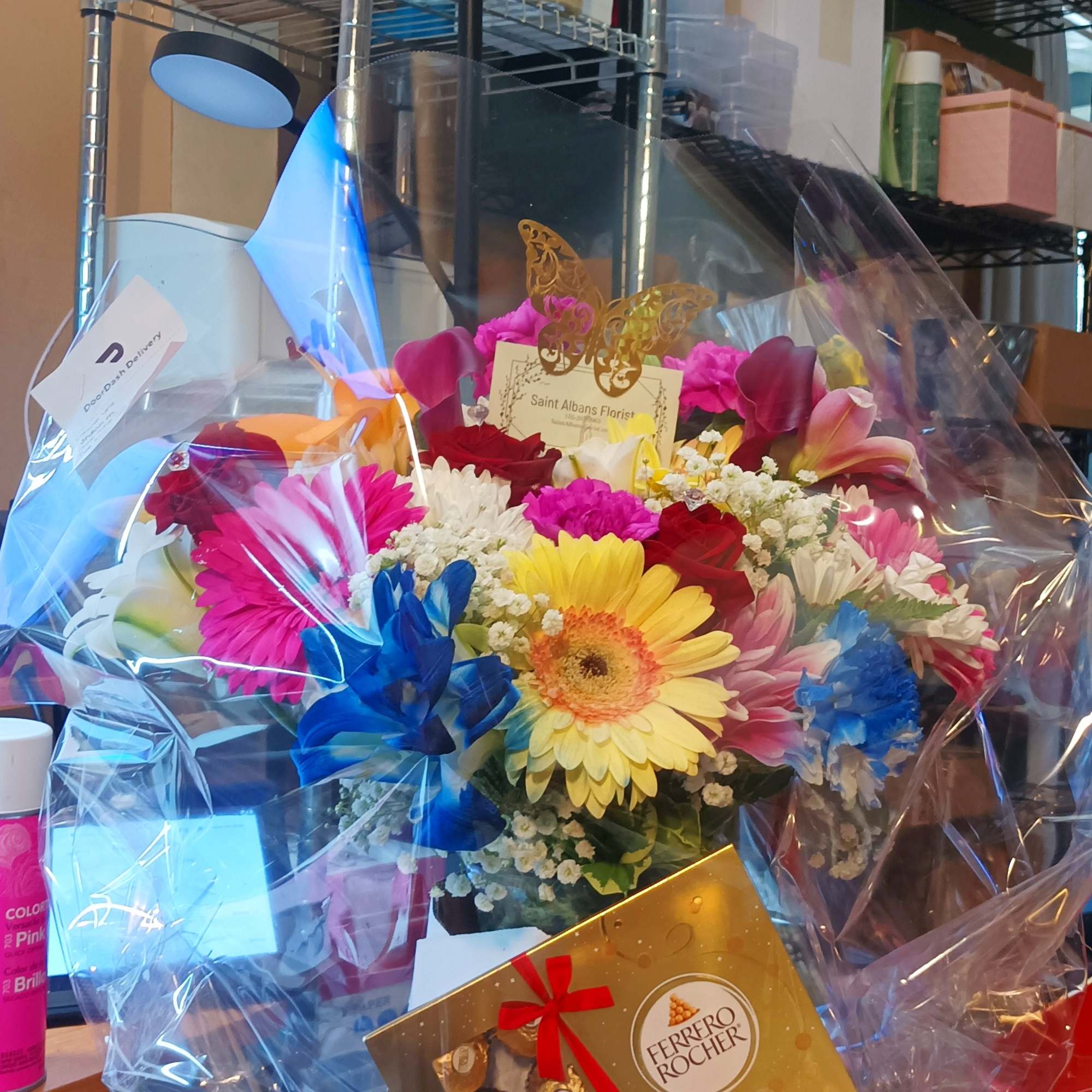 Mixed bouquet with gerbera daisies, roses, and lilies in clear wrap beside chocolates
