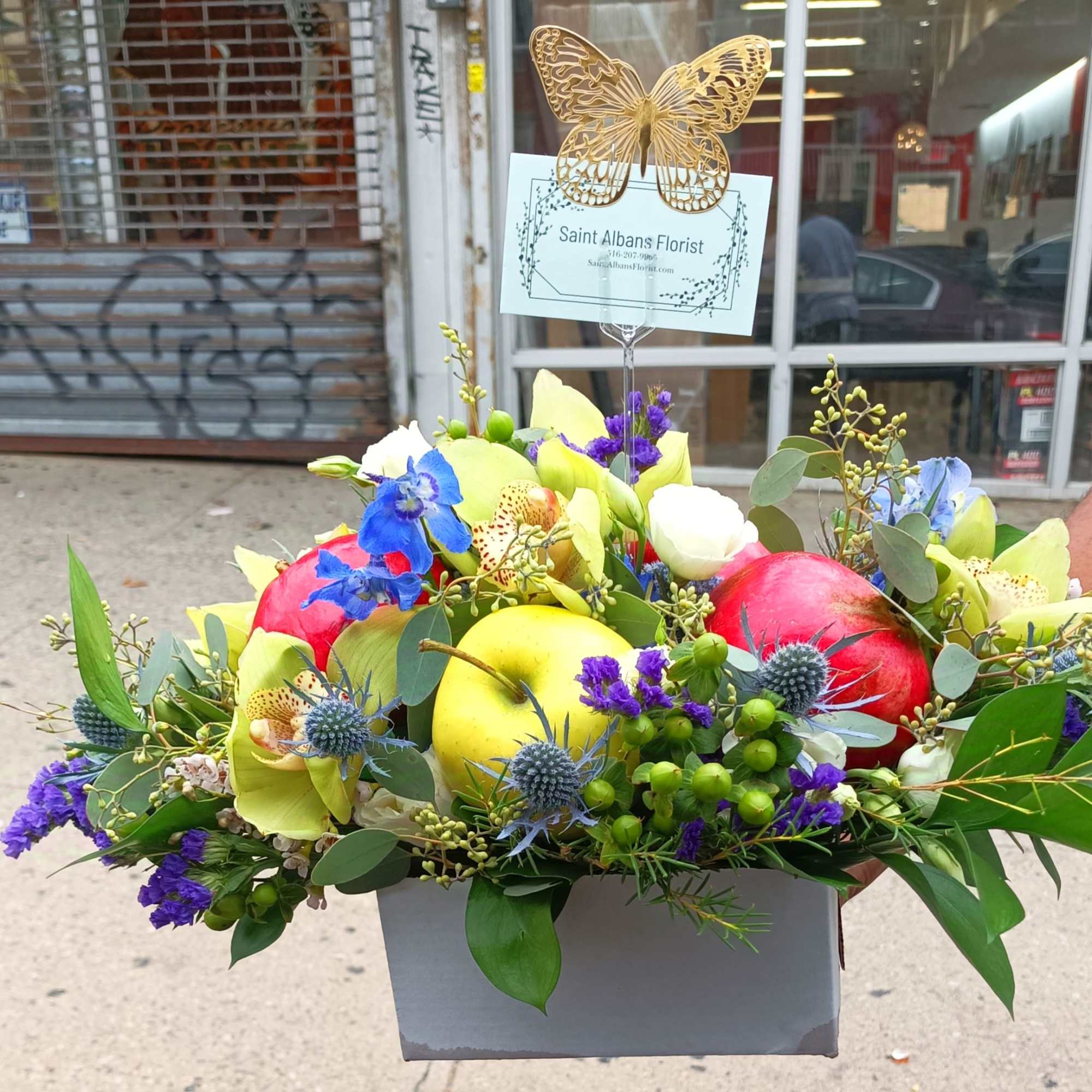 Colorful fruit-and-flower arrangement in a gray box with a butterfly topper