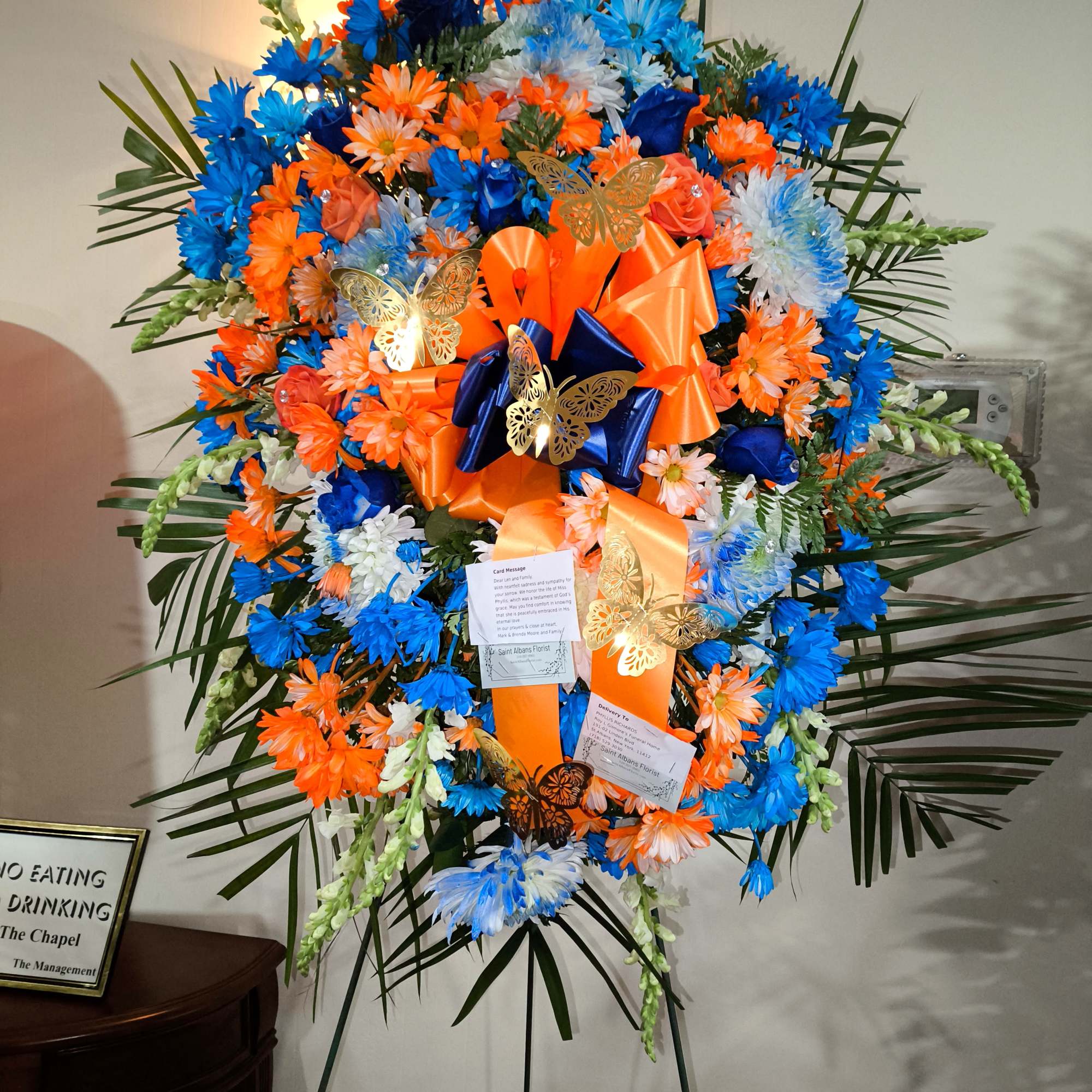 Large standing floral spray in blue, orange, and white with ribbons and butterflies