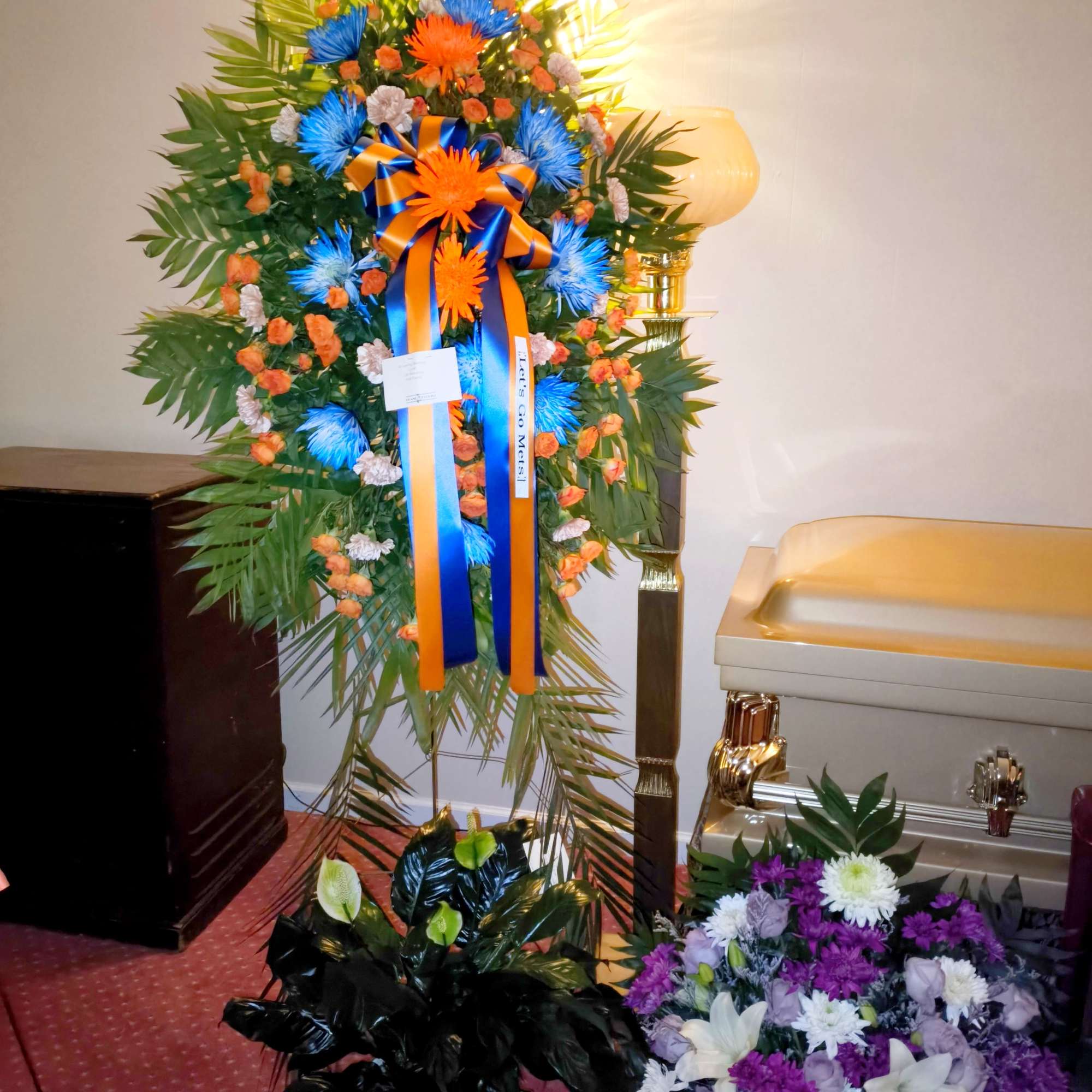 Standing funeral wreath with orange and blue flowers and ribbon beside a casket