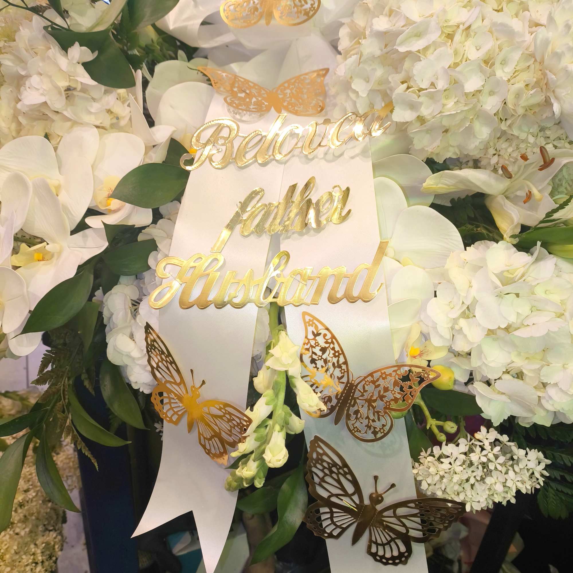White floral tribute with gold butterfly accents and a "Beloved father husband" sign