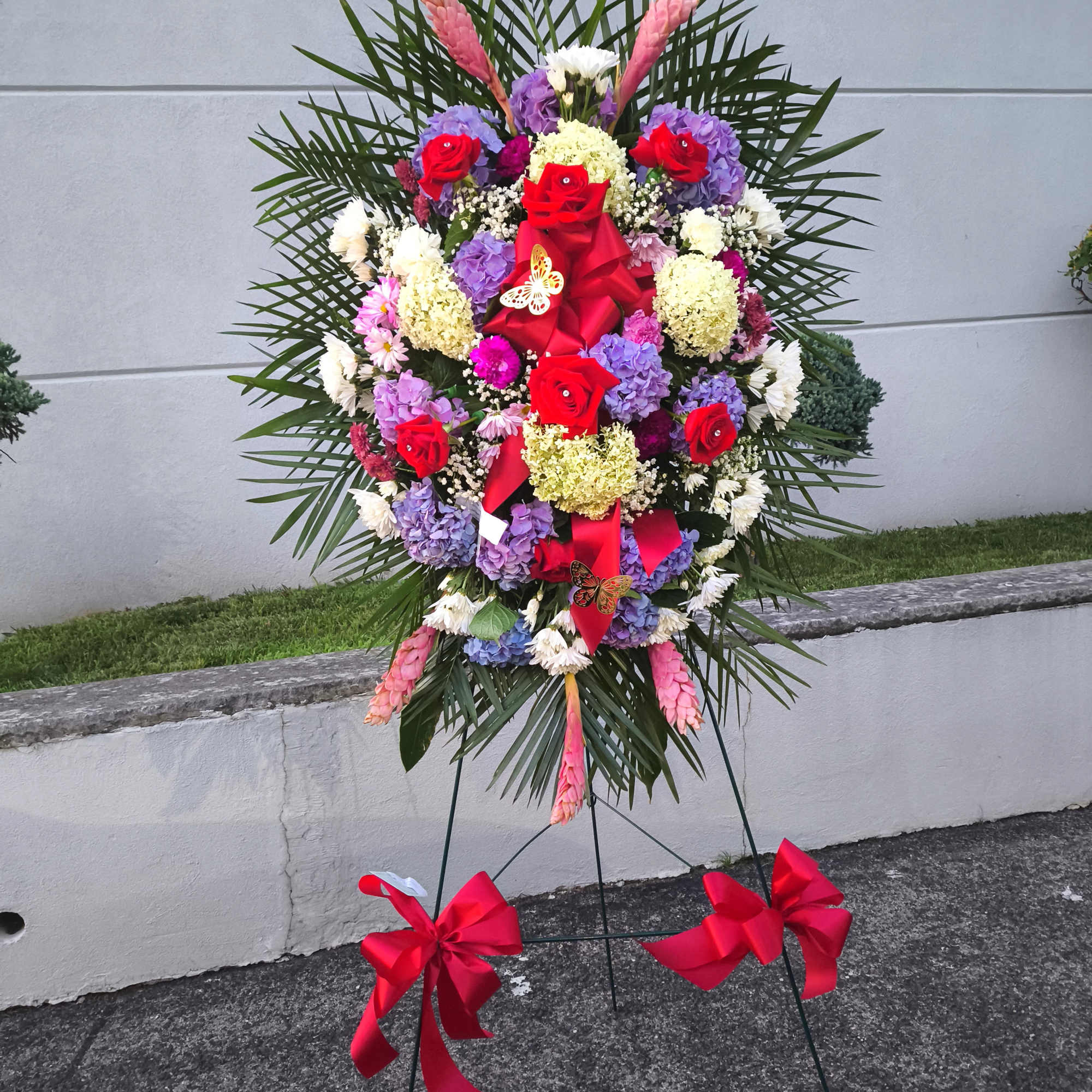 Standing floral wreath with red roses, purple hydrangeas, and pink ribbons