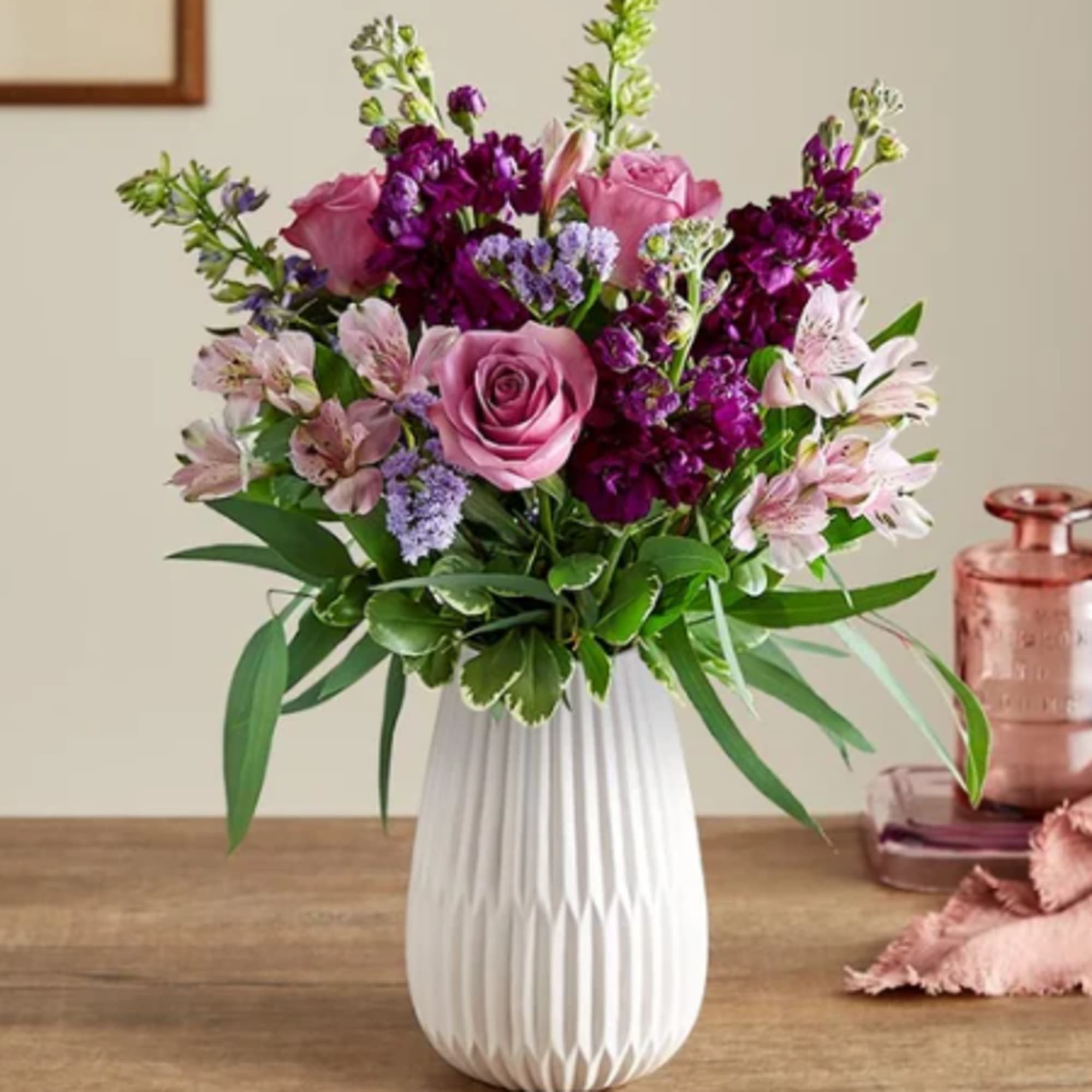 Mixed bouquet of pink roses and purple flowers in a white textured ceramic vase on a wooden table