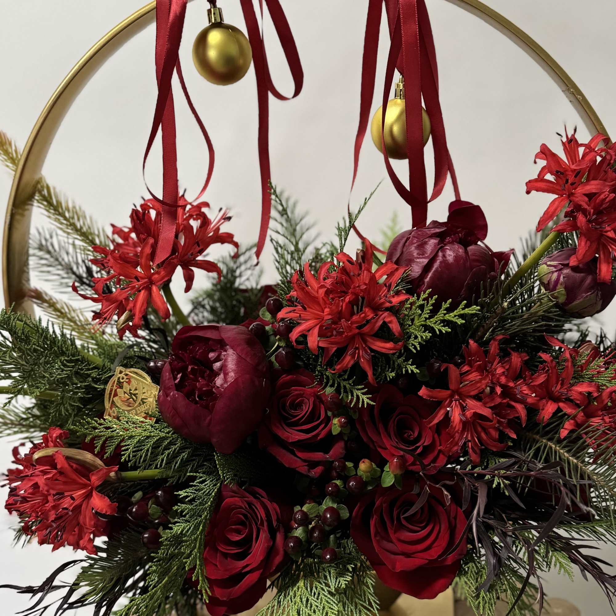 Red roses and burgundy blooms arranged in a gold hoop with ribbon and ornaments.