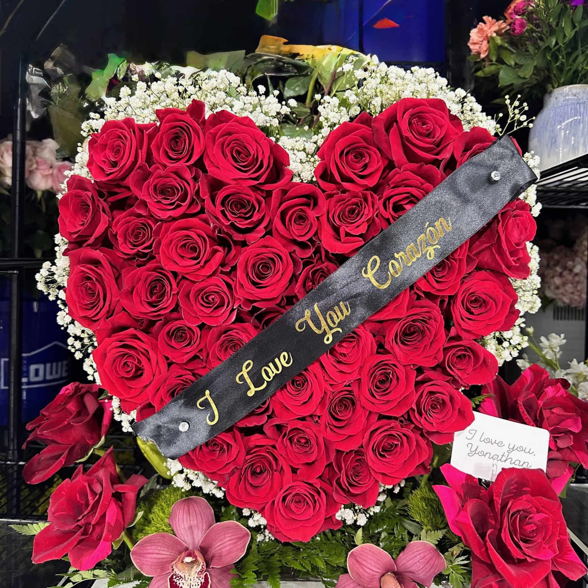 Heart-shaped red rose arrangement with a black ribbon and orchids