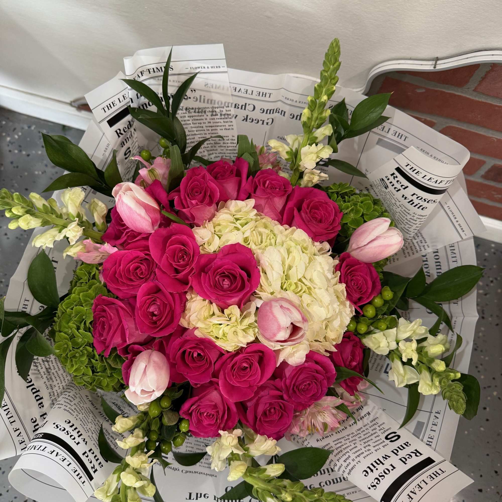 Bouquet of hot pink roses, pale tulips, and white hydrangea wrapped in newspaper