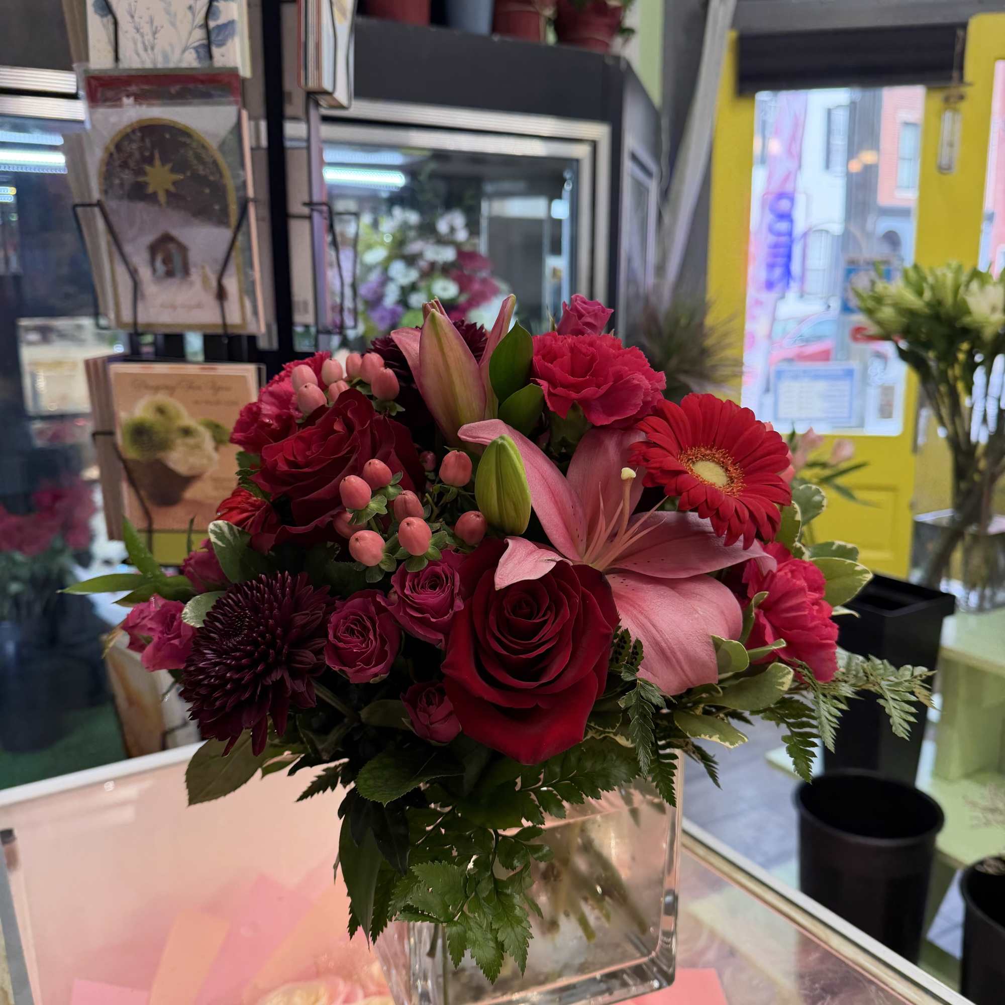 Mixed bouquet of red, pink, and burgundy flowers in a clear square vase
