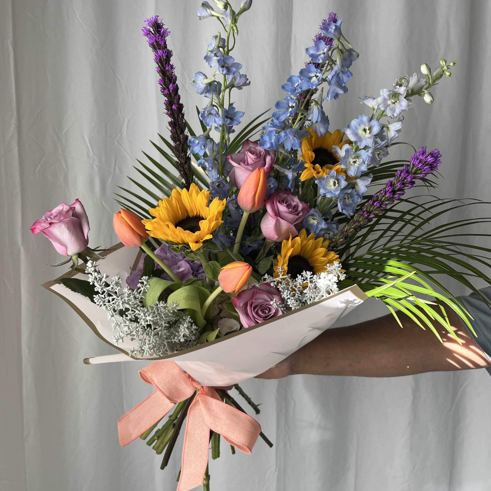 Bouquet of sunflowers, roses, tulips, and blue delphinium in white wrap