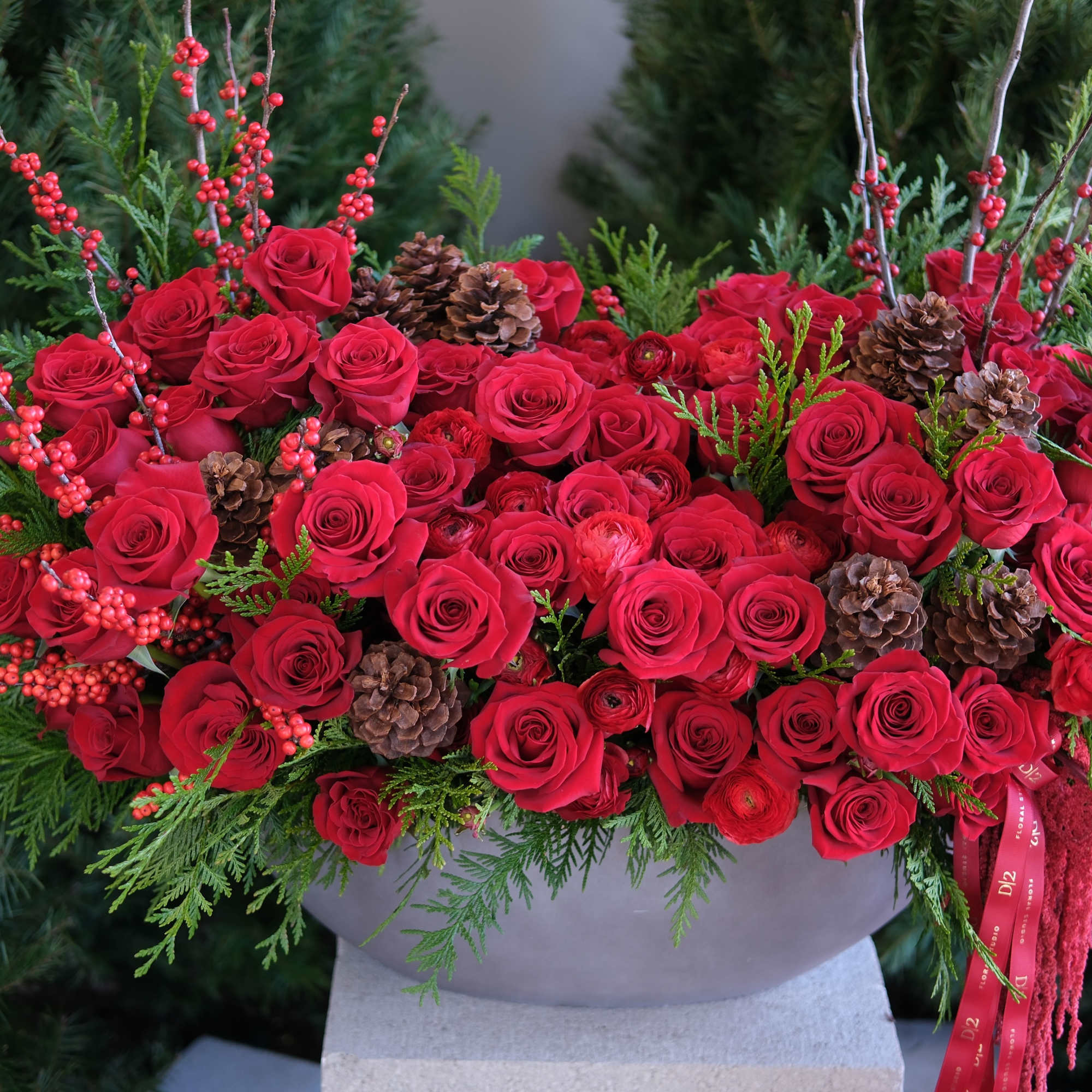 Red rose arrangement with pinecones and evergreen branches in a gray container