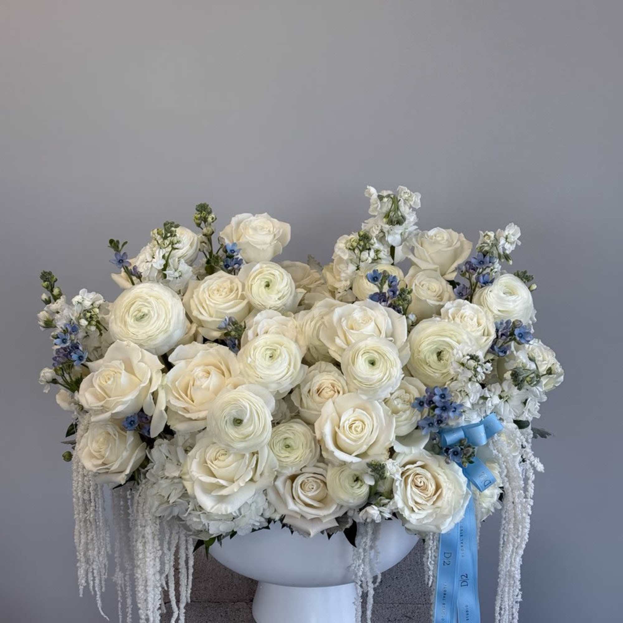 White rose and ranunculus arrangement in a white pedestal vase with blue ribbon