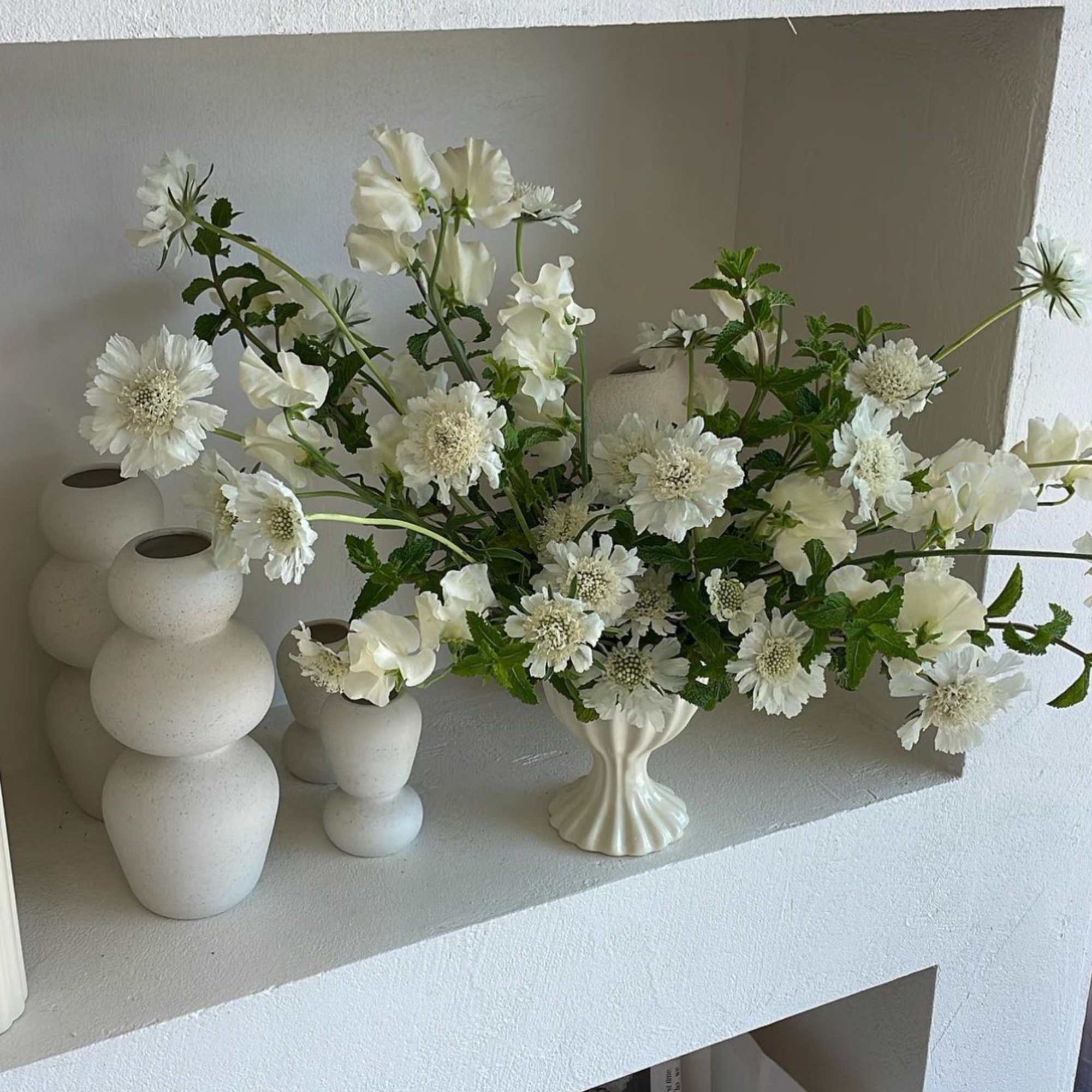 White floral arrangement in a cream vase with several white ceramic vases nearby