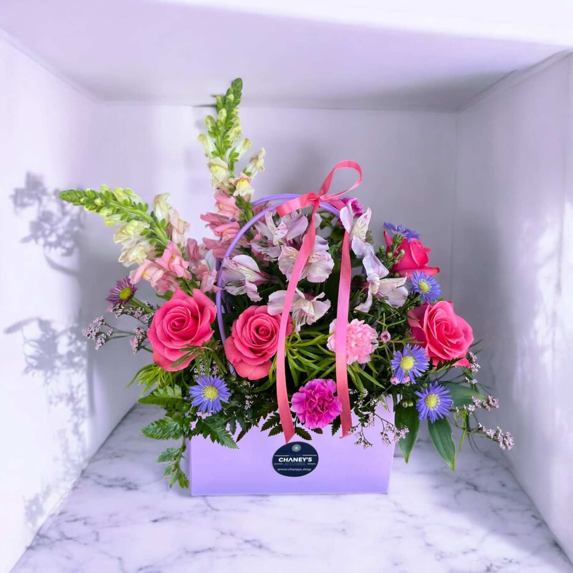 Pink roses and mixed flowers arranged in a lavender gift box with ribbon.