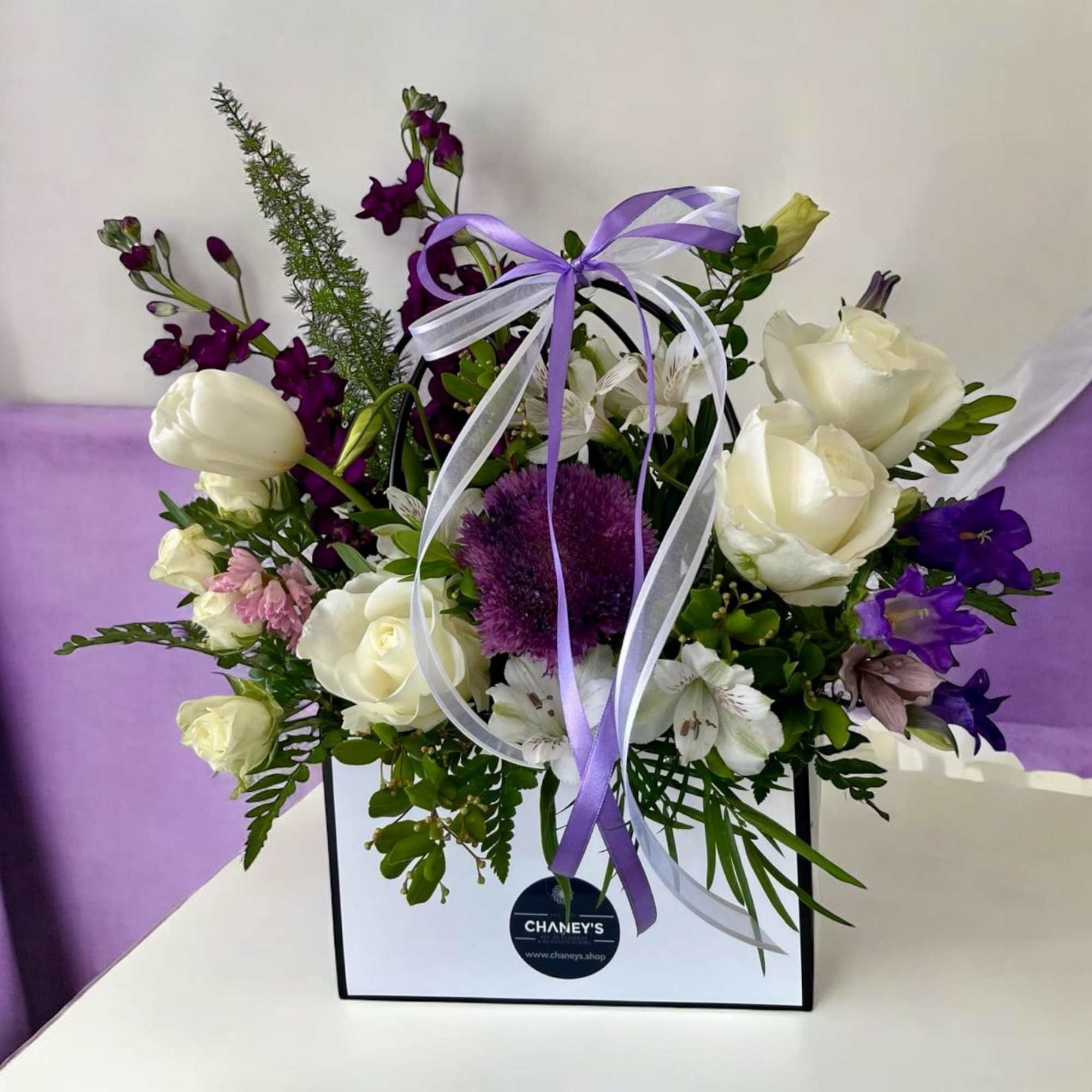 Purple and white floral arrangement in a gift bag with ribbon