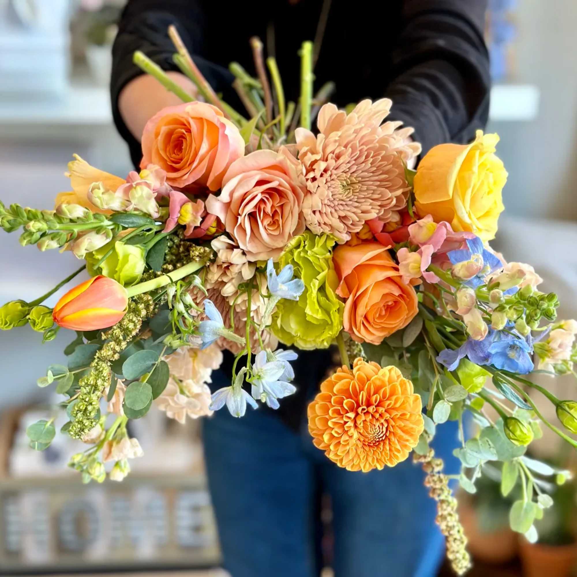 Handheld bouquet of peach, yellow, and orange flowers with blue accents