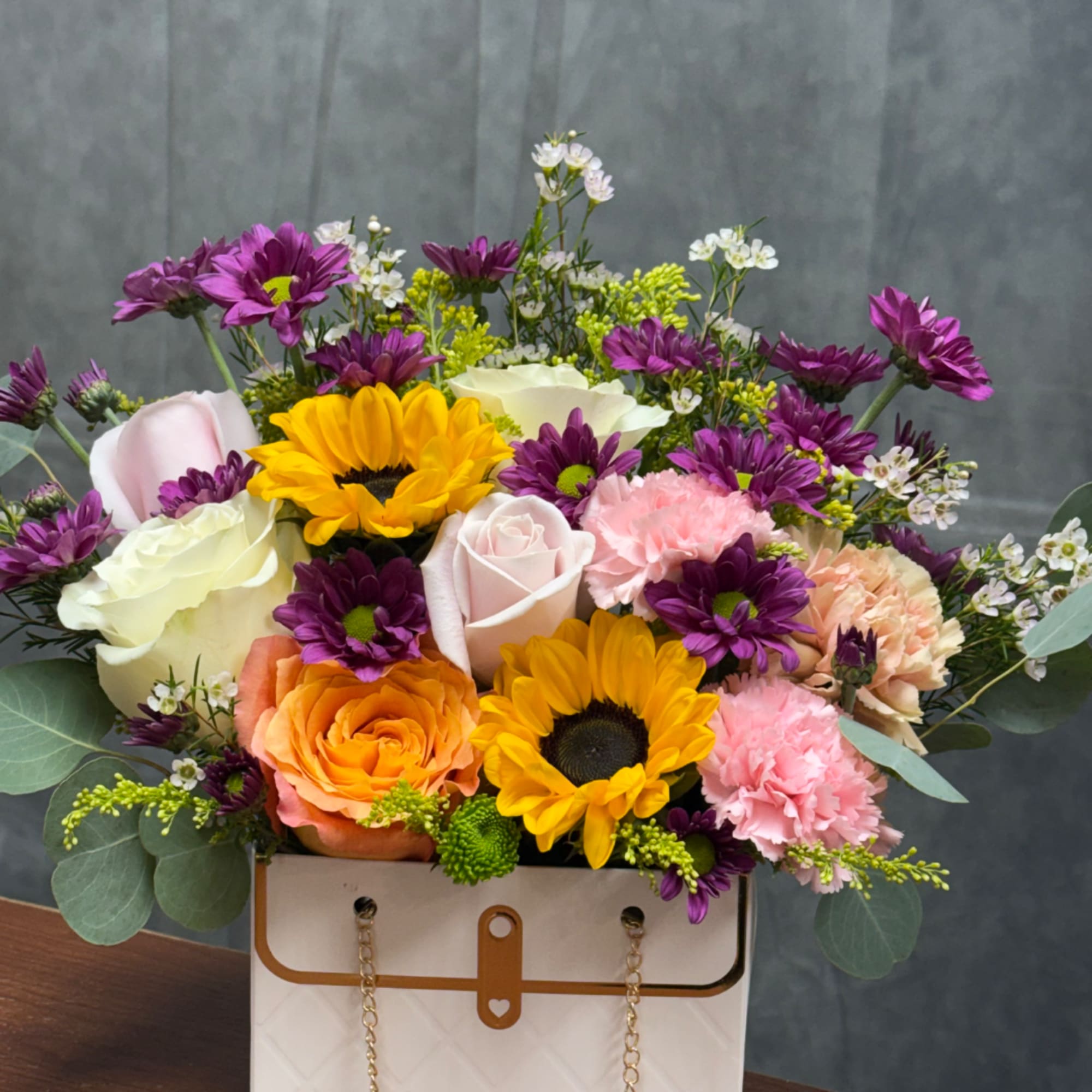 Mixed bouquet with roses, sunflowers, carnations, and purple daisies in a white box bag