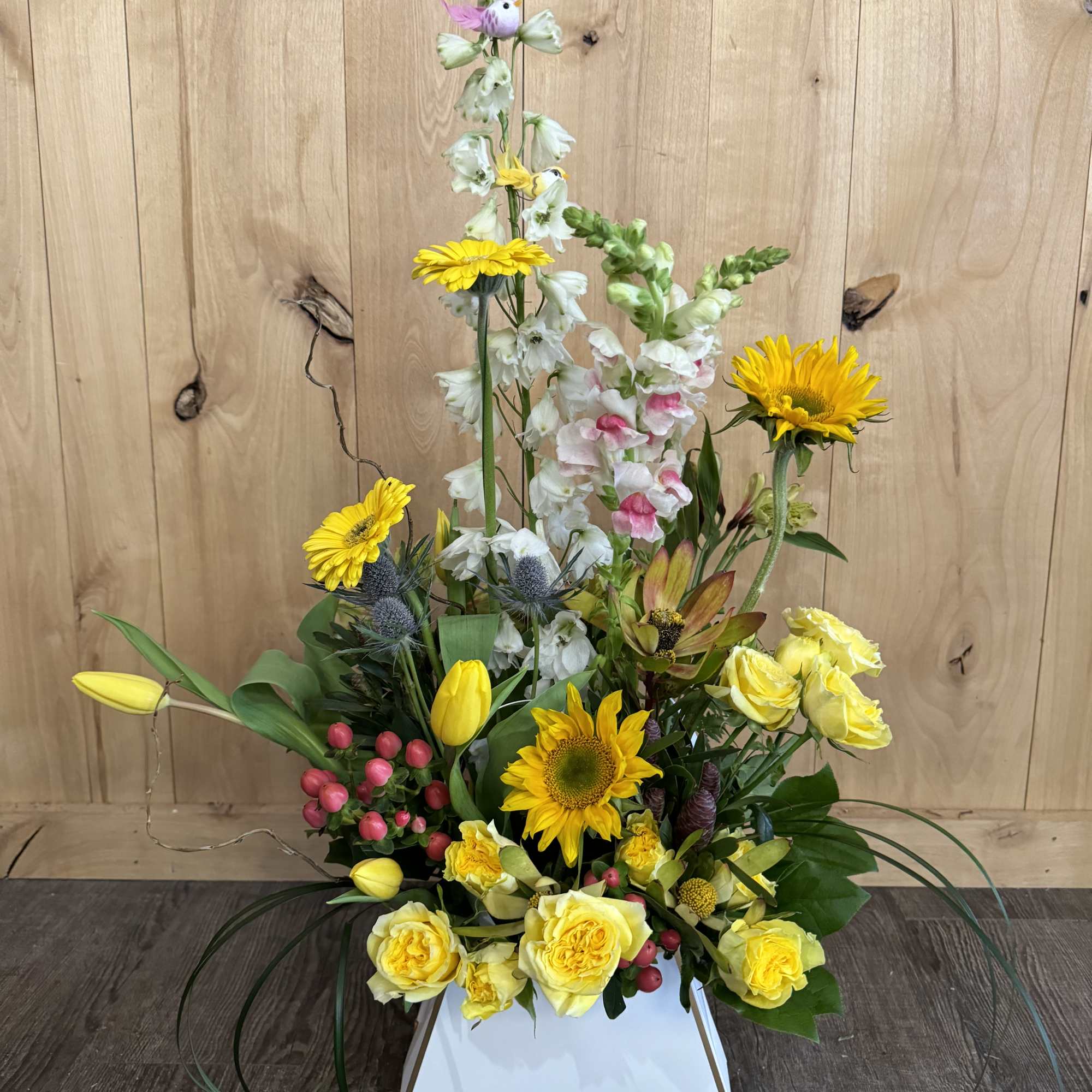 Tall floral arrangement with yellow roses, sunflowers, and white snapdragons in a white box