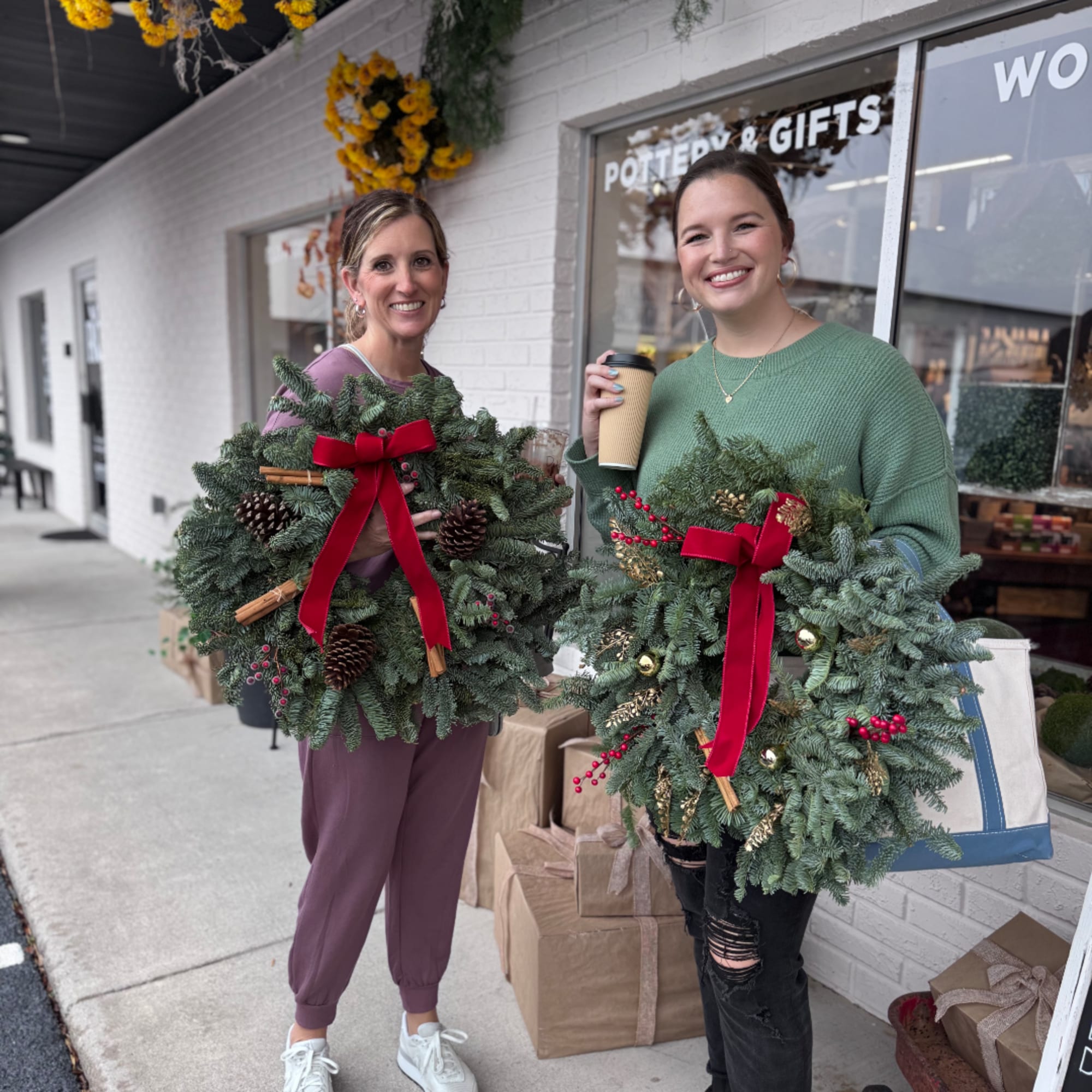 Two women hold evergreen wreaths with red bows and pinecones outside a shop.