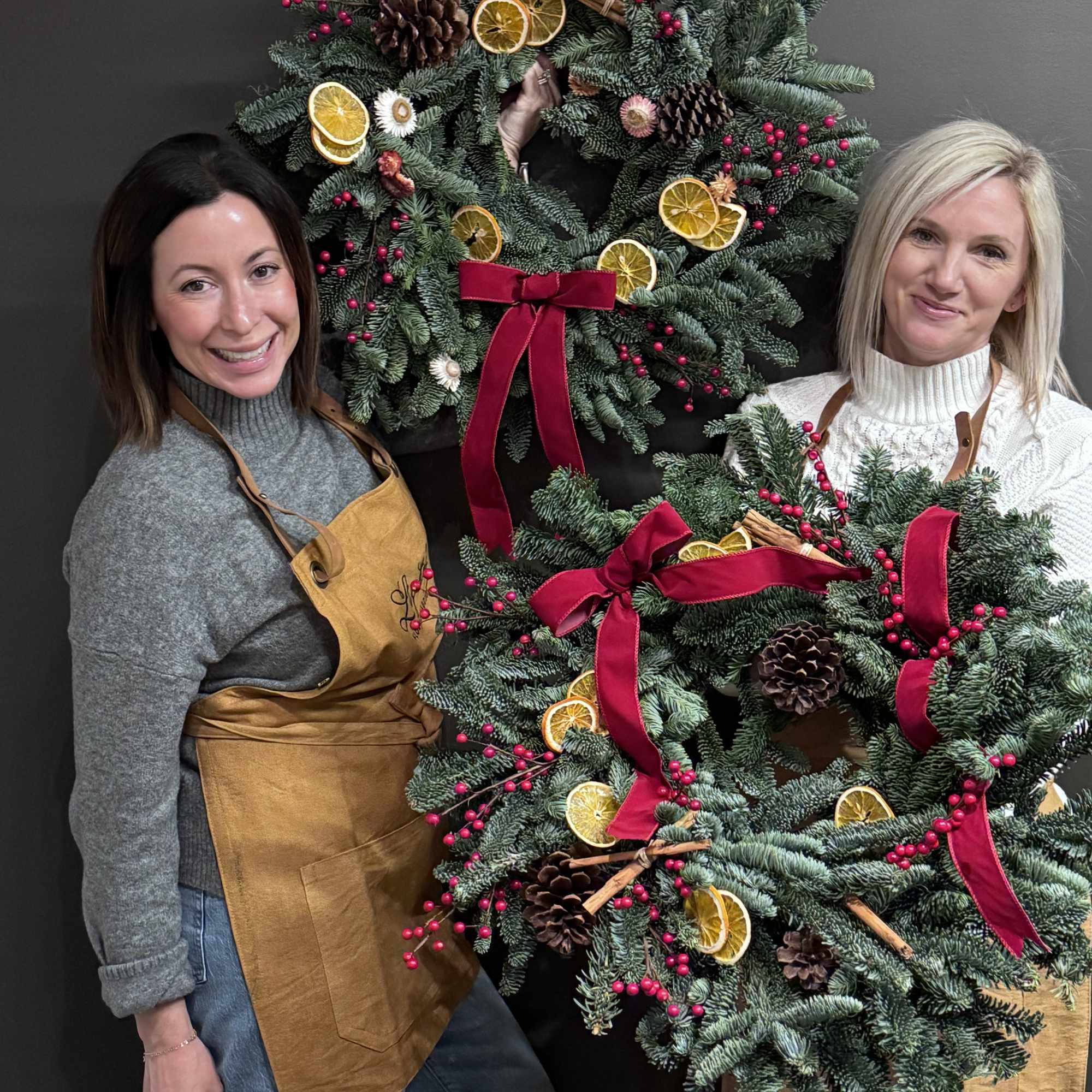 Two women hold decorated evergreen wreaths with red bows and dried citrus slices.