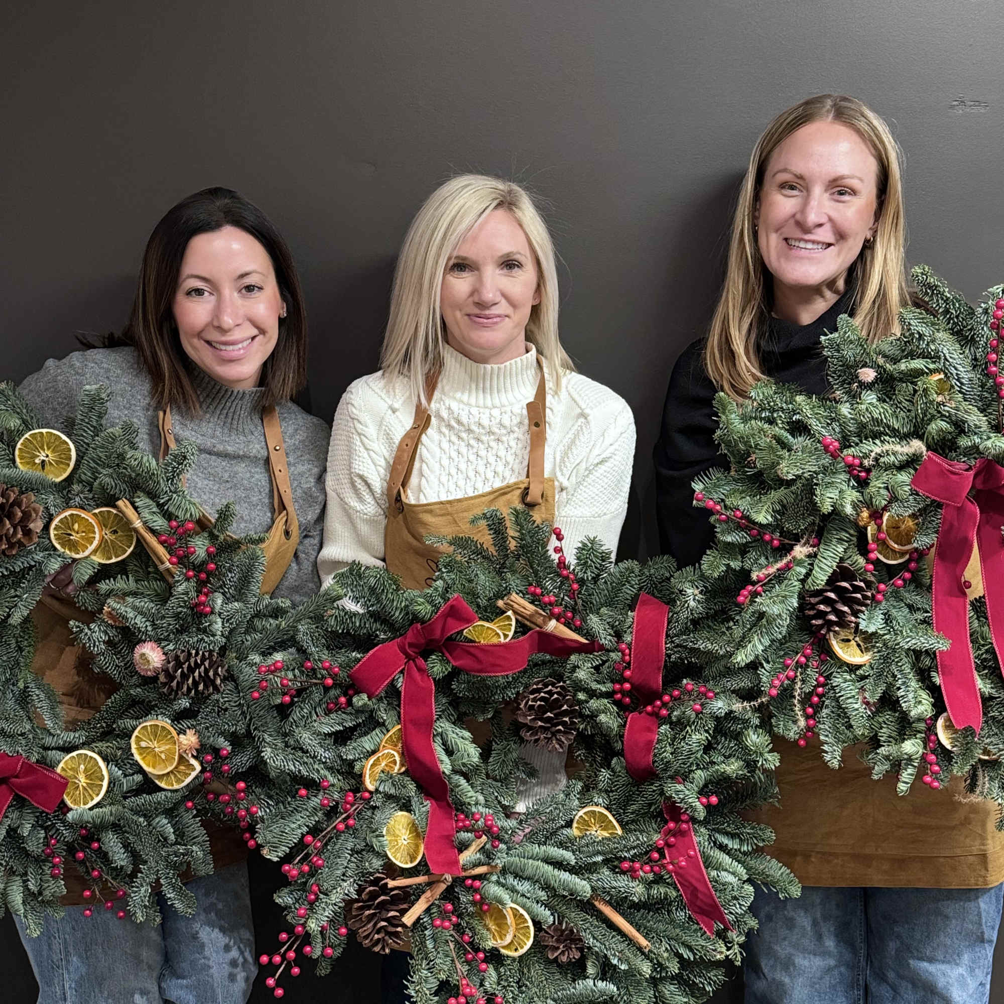 Three women hold large holiday wreaths with red ribbons and dried citrus.