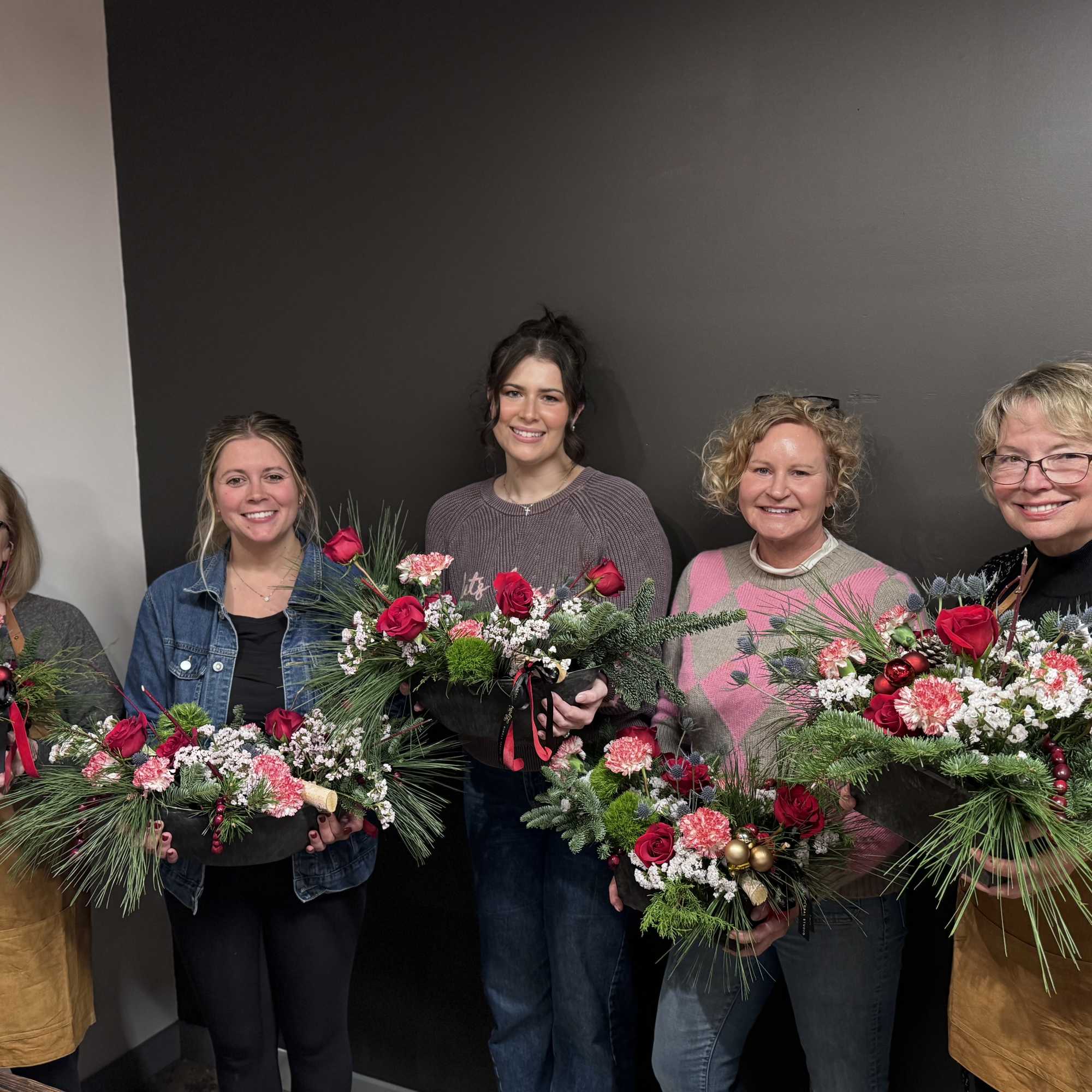 Five women hold holiday floral arrangements with red roses and pine accents.
