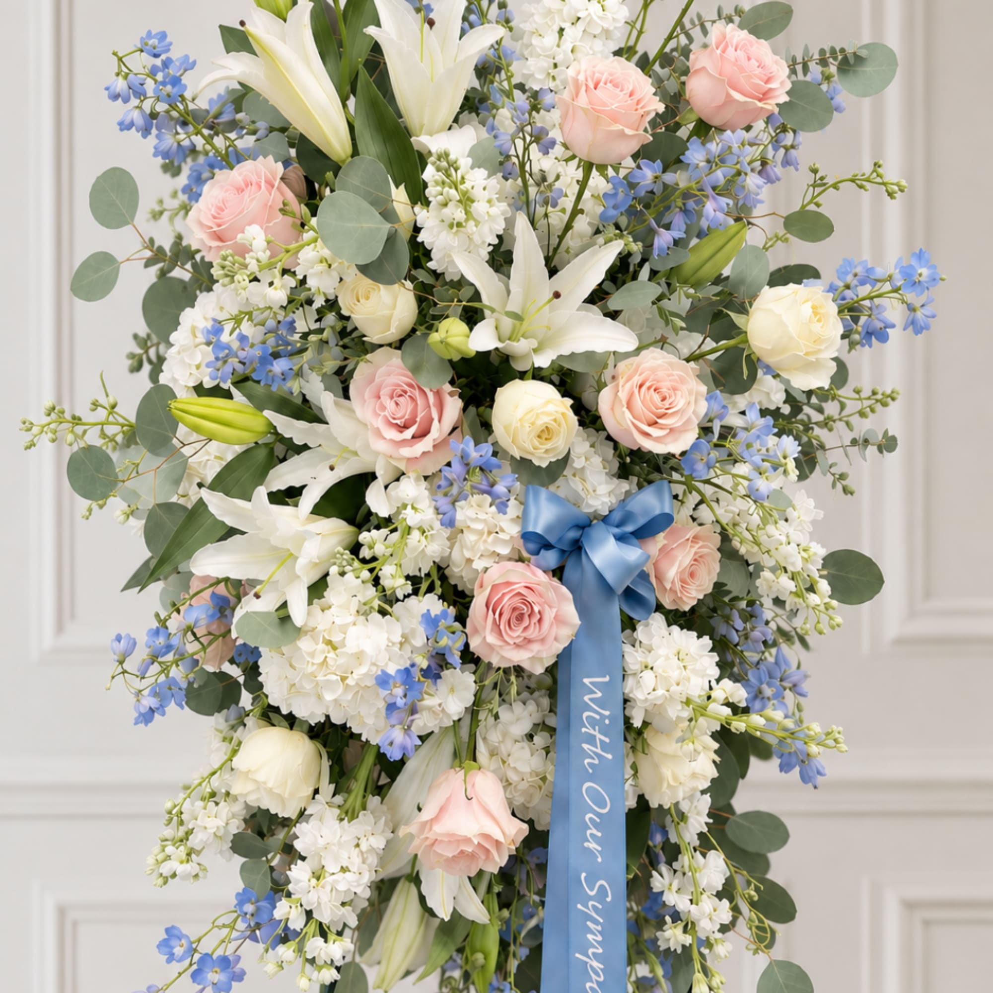 Tall pastel floral spray with roses, lilies, and blue flowers, finished with a blue ribbon