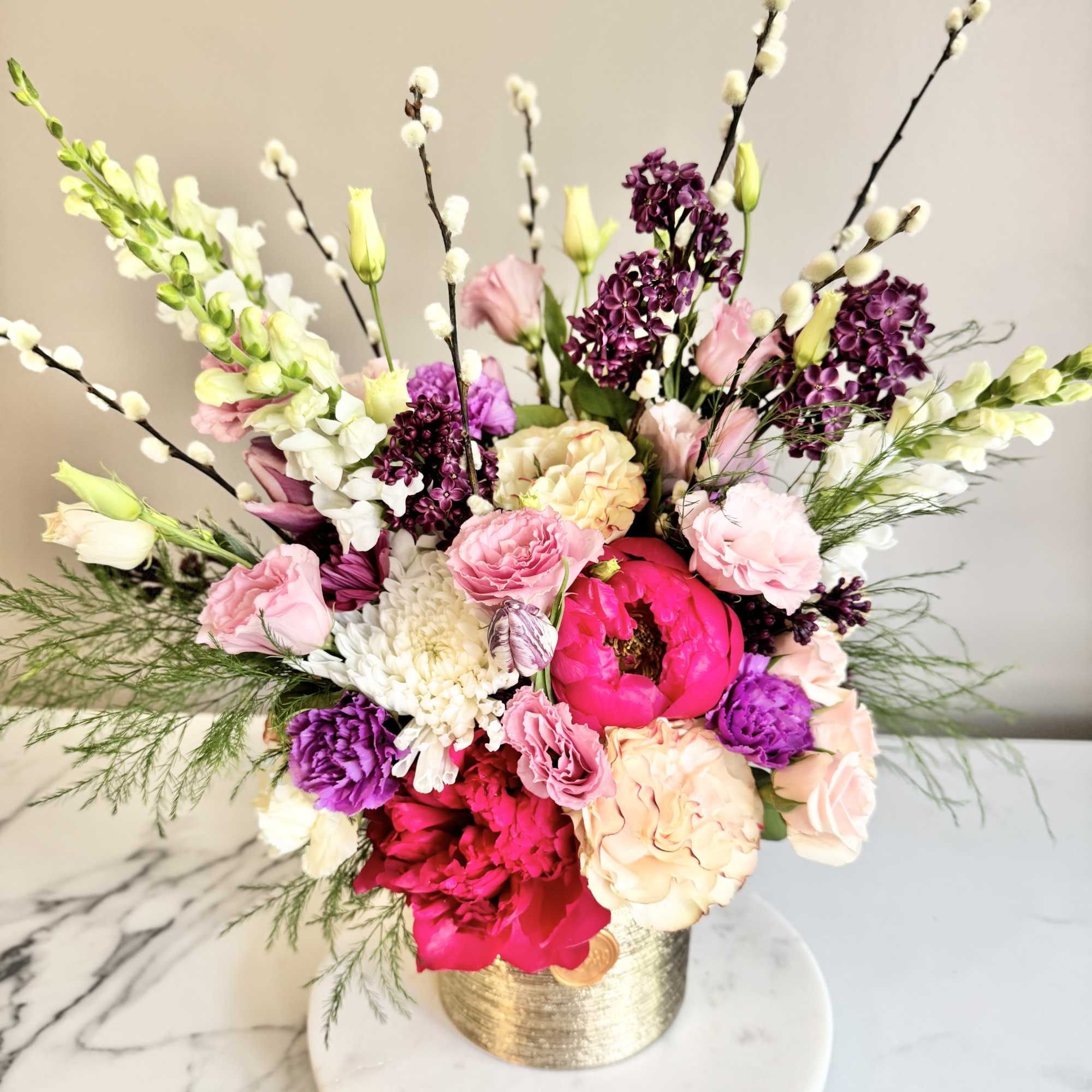 Lush arrangement of pink, peach, purple and white flowers in a gold vase on a marble surface