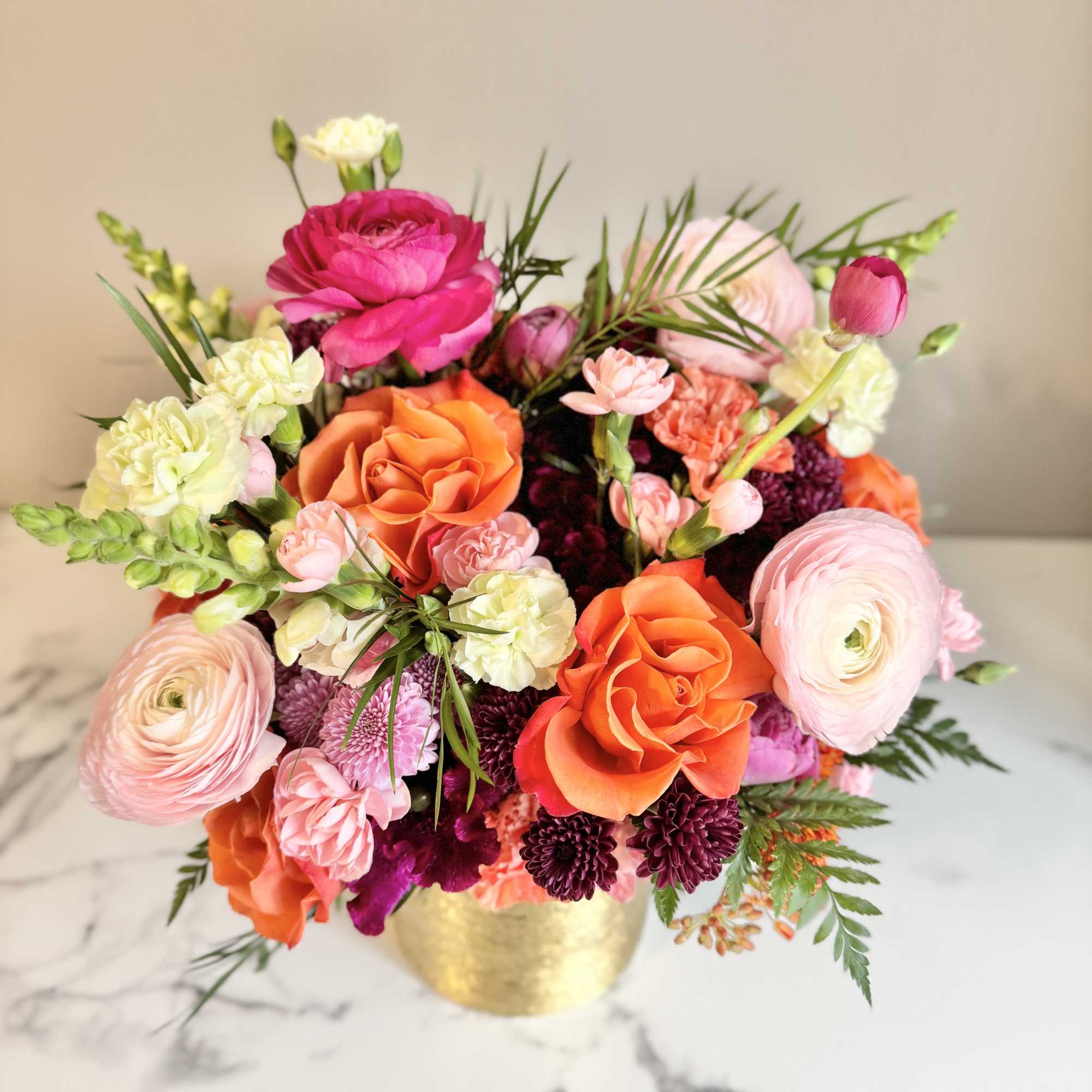 Bright arrangement of pink ranunculus, orange roses, and mixed blooms in a gold vase on a marble surface