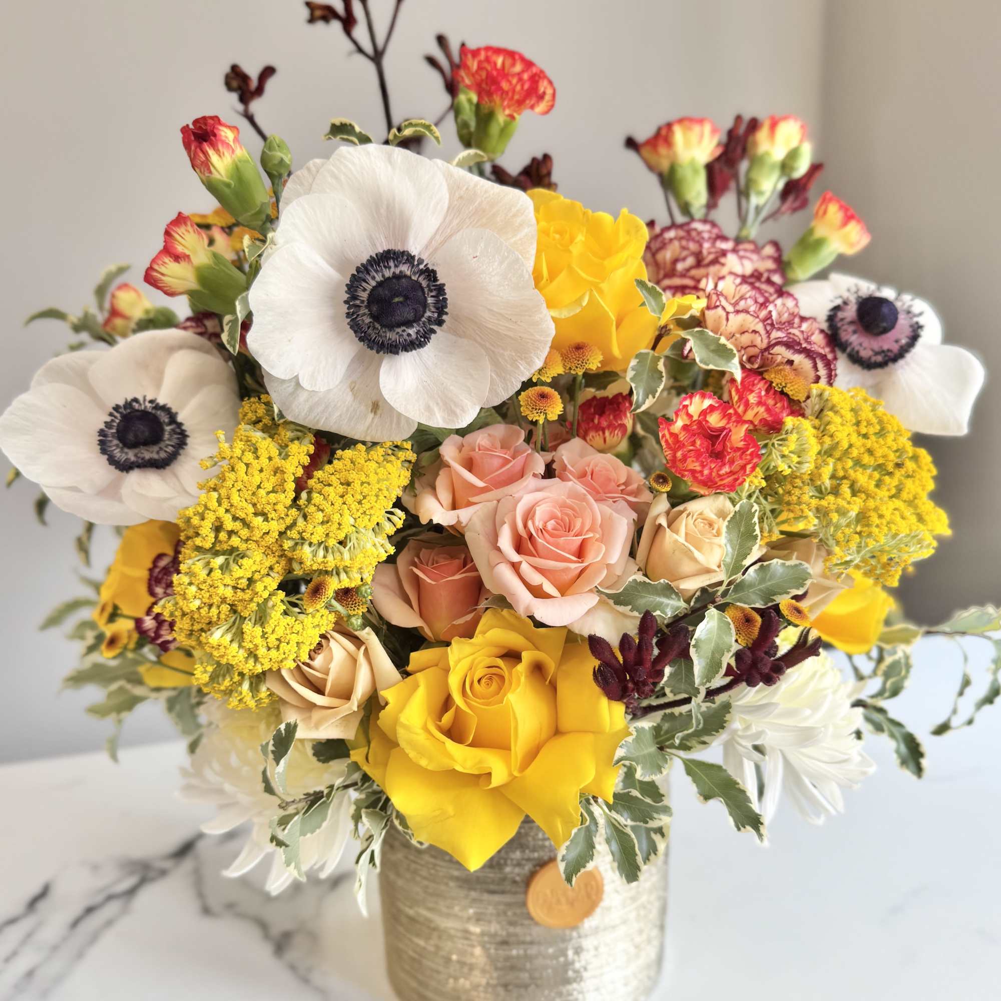 Bright mixed arrangement with white anemones, yellow and peach roses, and carnations in a metallic vase