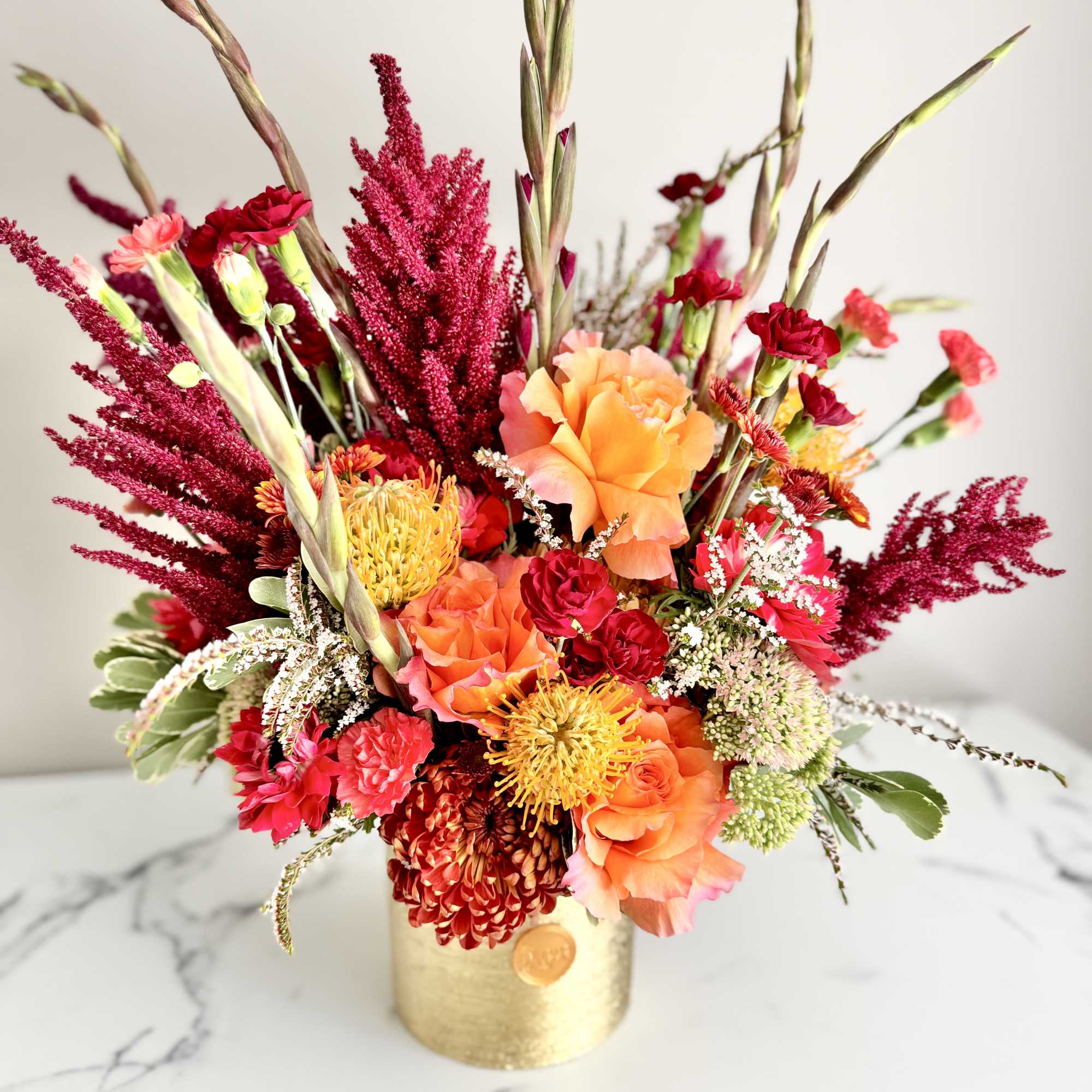 Vibrant arrangement of orange roses, red carnations and yellow protea in a gold cylinder vase
