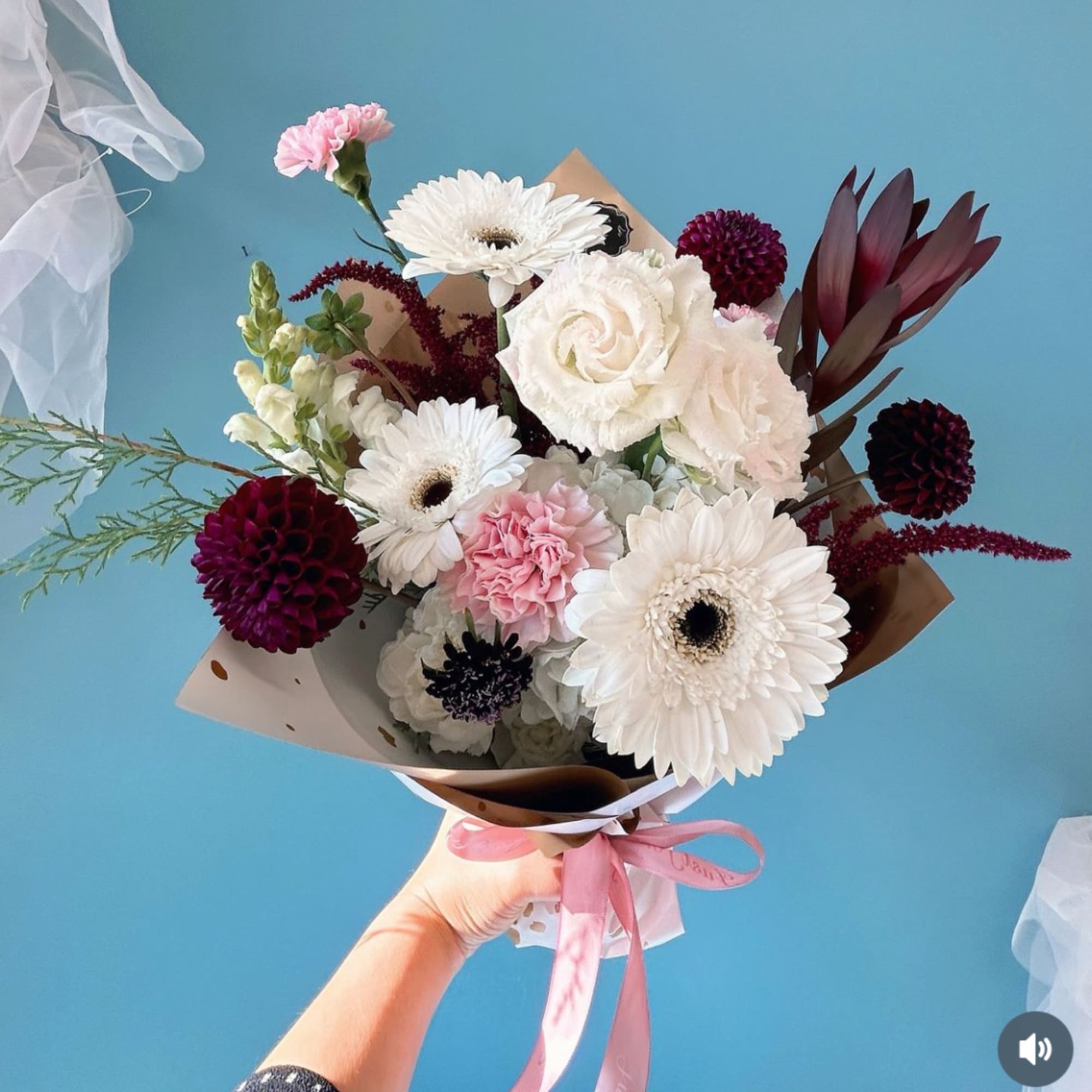 Handheld bouquet of white, pink, and burgundy flowers wrapped in paper with a pink ribbon