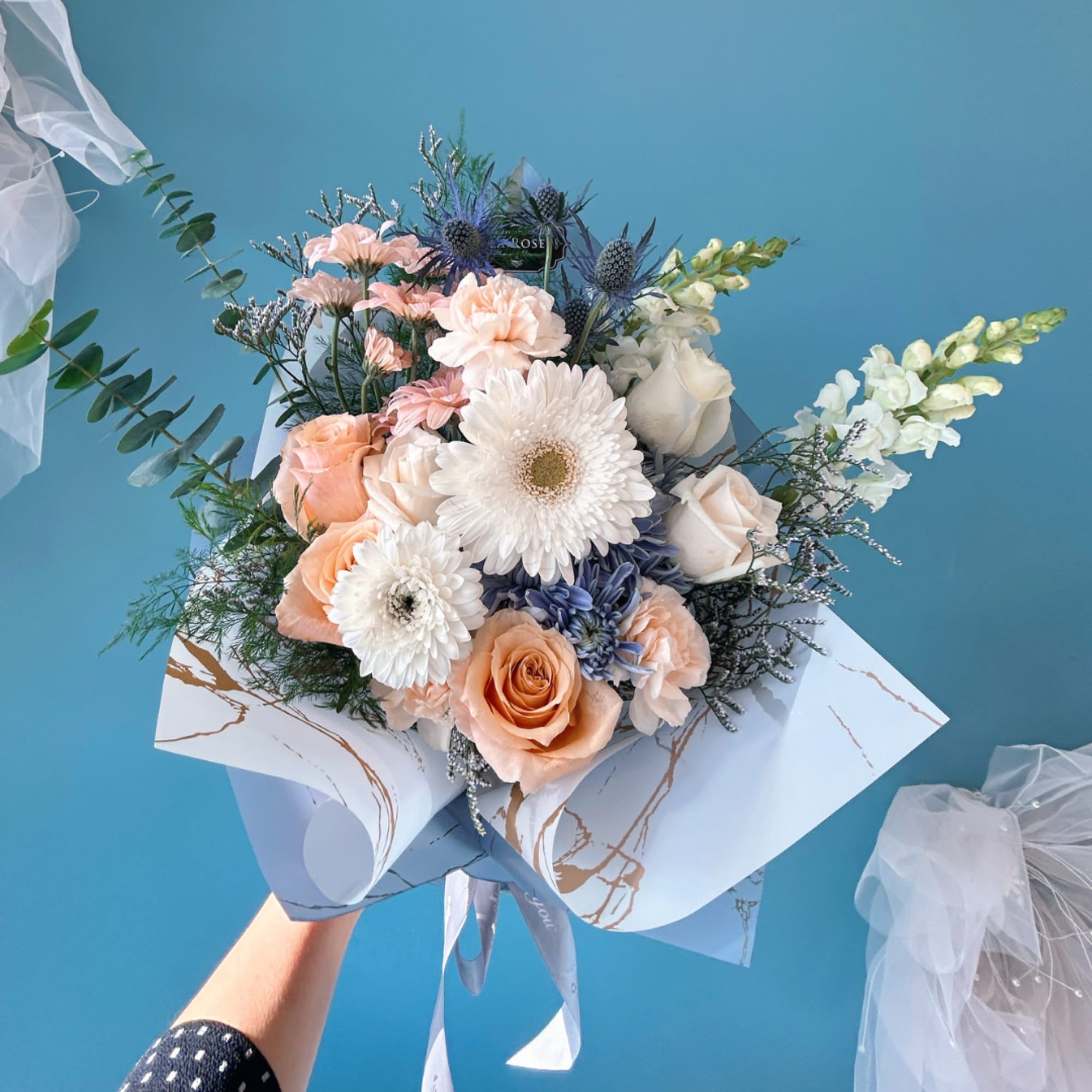 Handheld bouquet of peach, white, and blue flowers wrapped in white paper