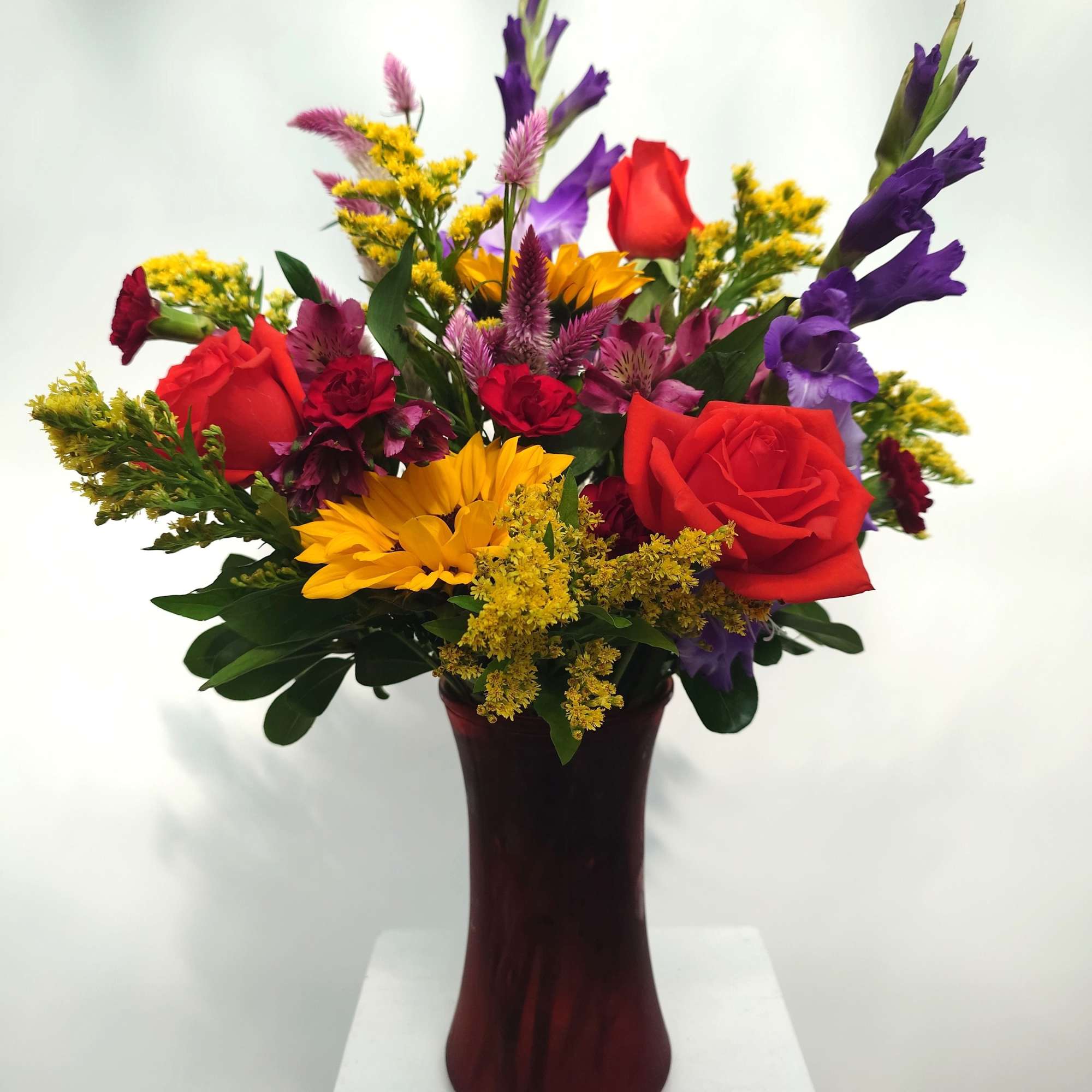 Mixed bouquet of red roses, yellow sunflowers, and purple flowers in a burgundy vase