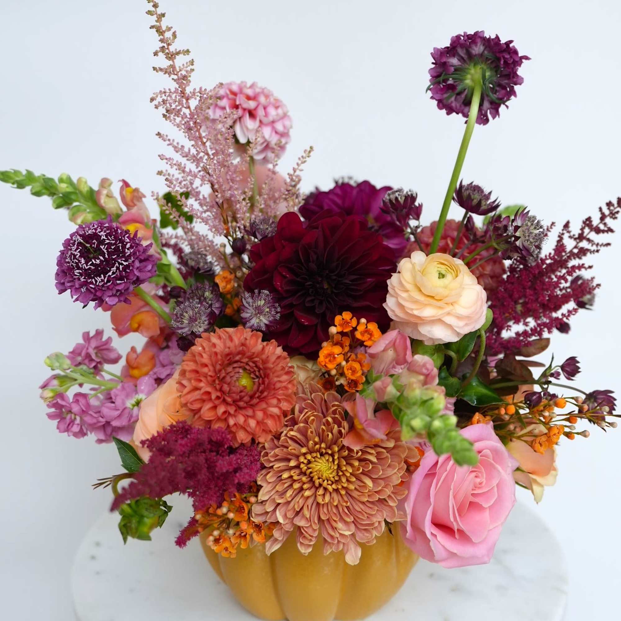 Mixed bouquet in a pumpkin-shaped vase with pink, purple, peach, and orange blooms.
