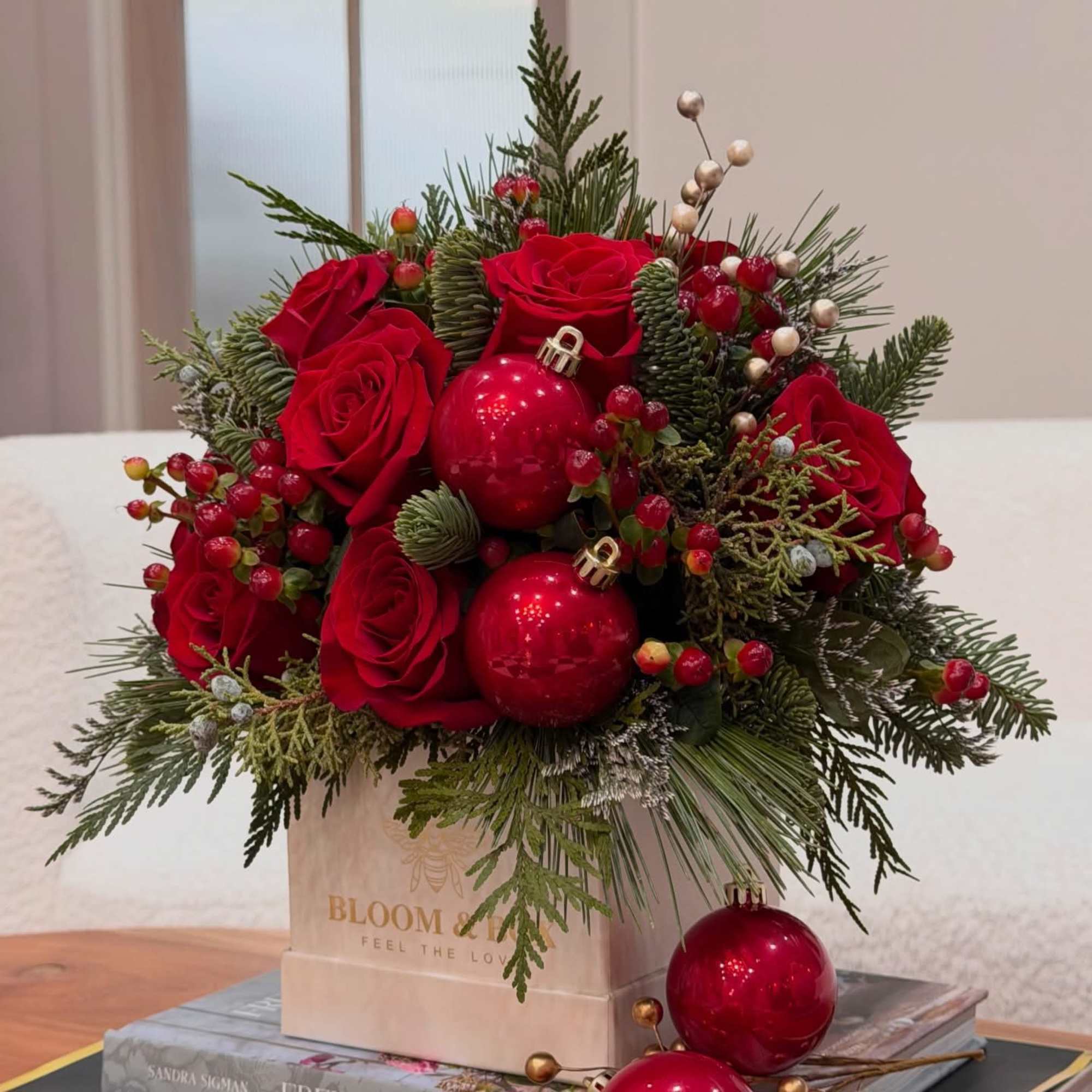 Red roses and holiday ornaments arranged in a gift box with evergreen accents