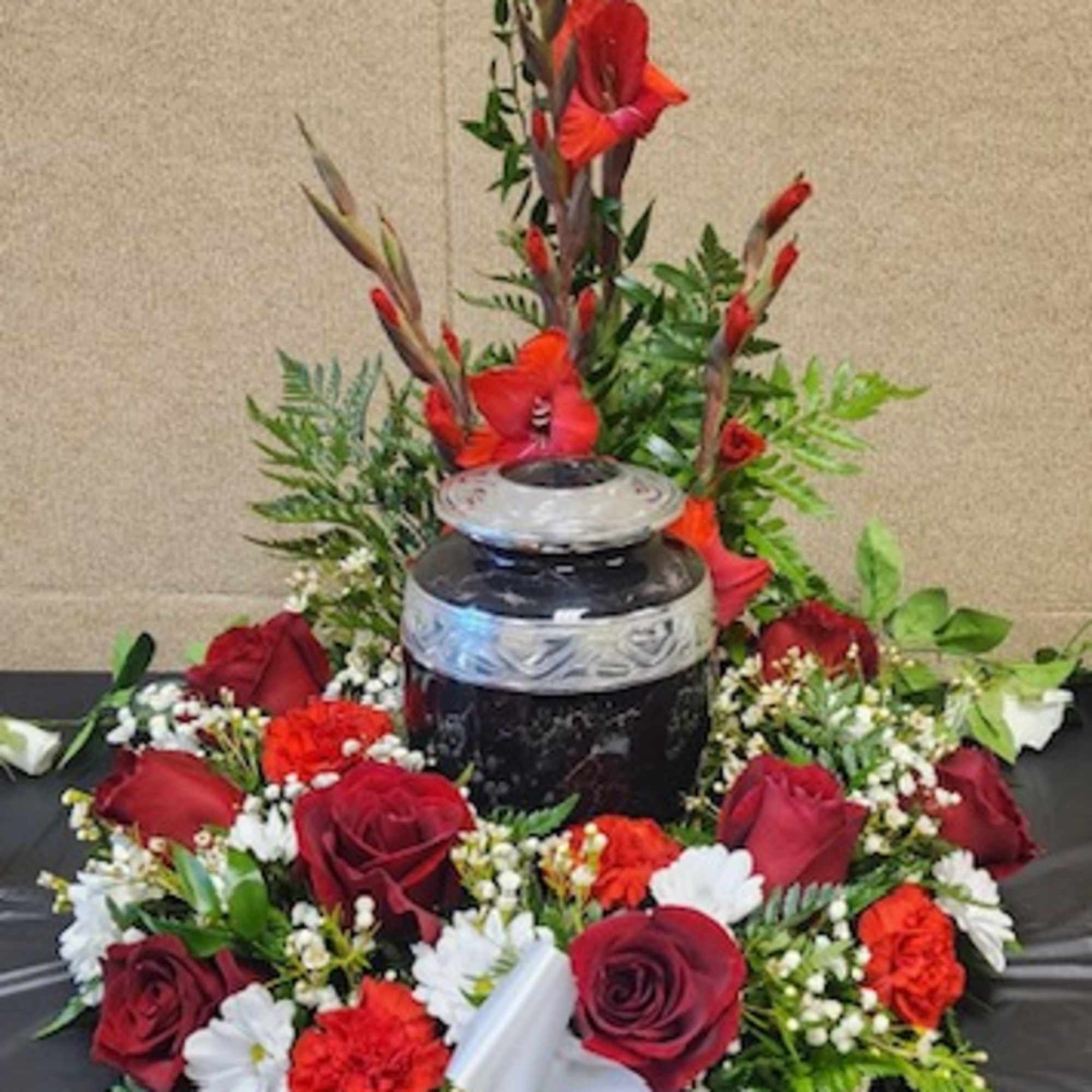 Red roses and carnations arranged around a black urn with a white ribbon.