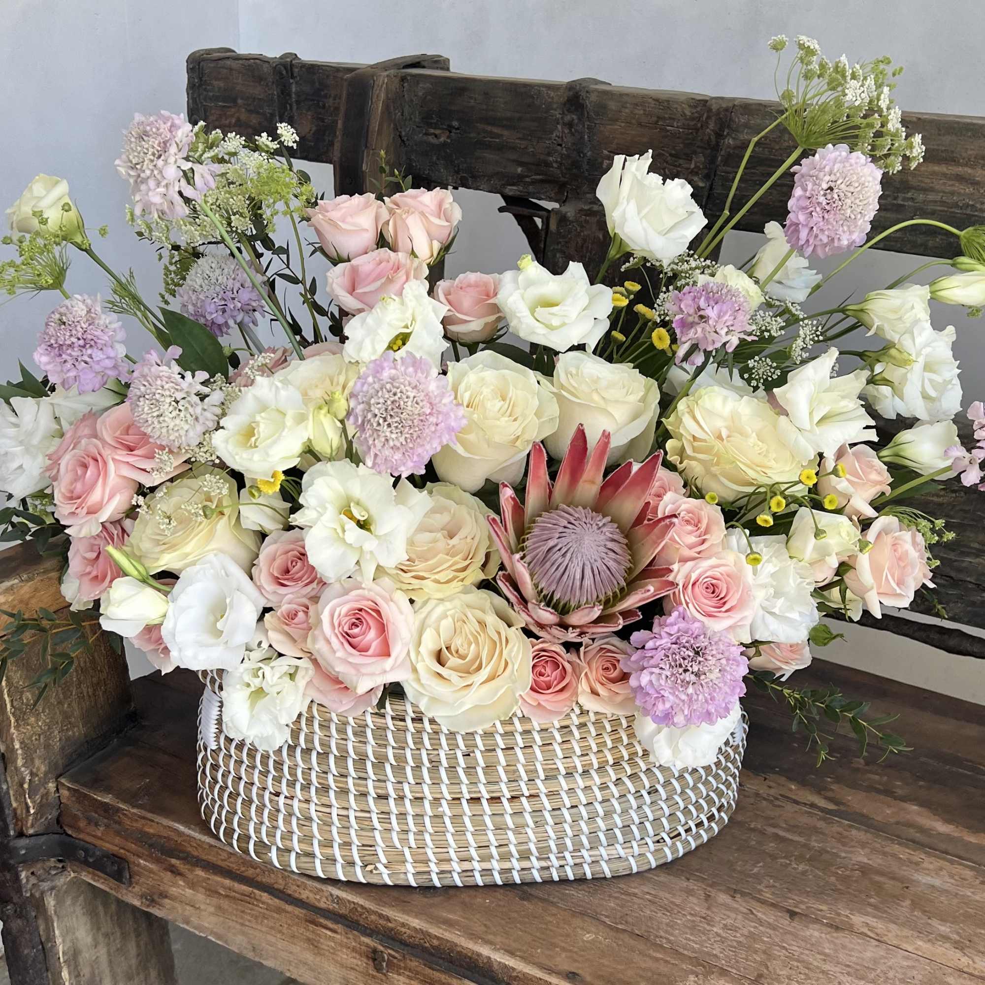 This flower basket features a striking protea with cream and soft pink