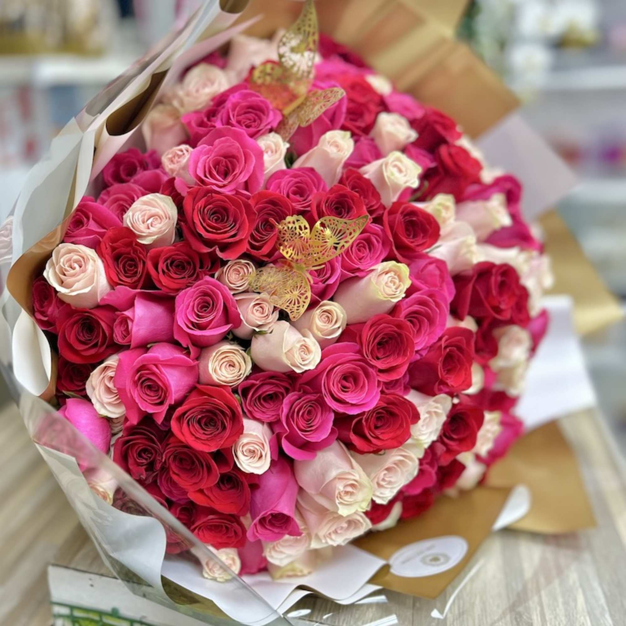 Large round bouquet of hot pink, red, and pale roses wrapped in white and gold paper