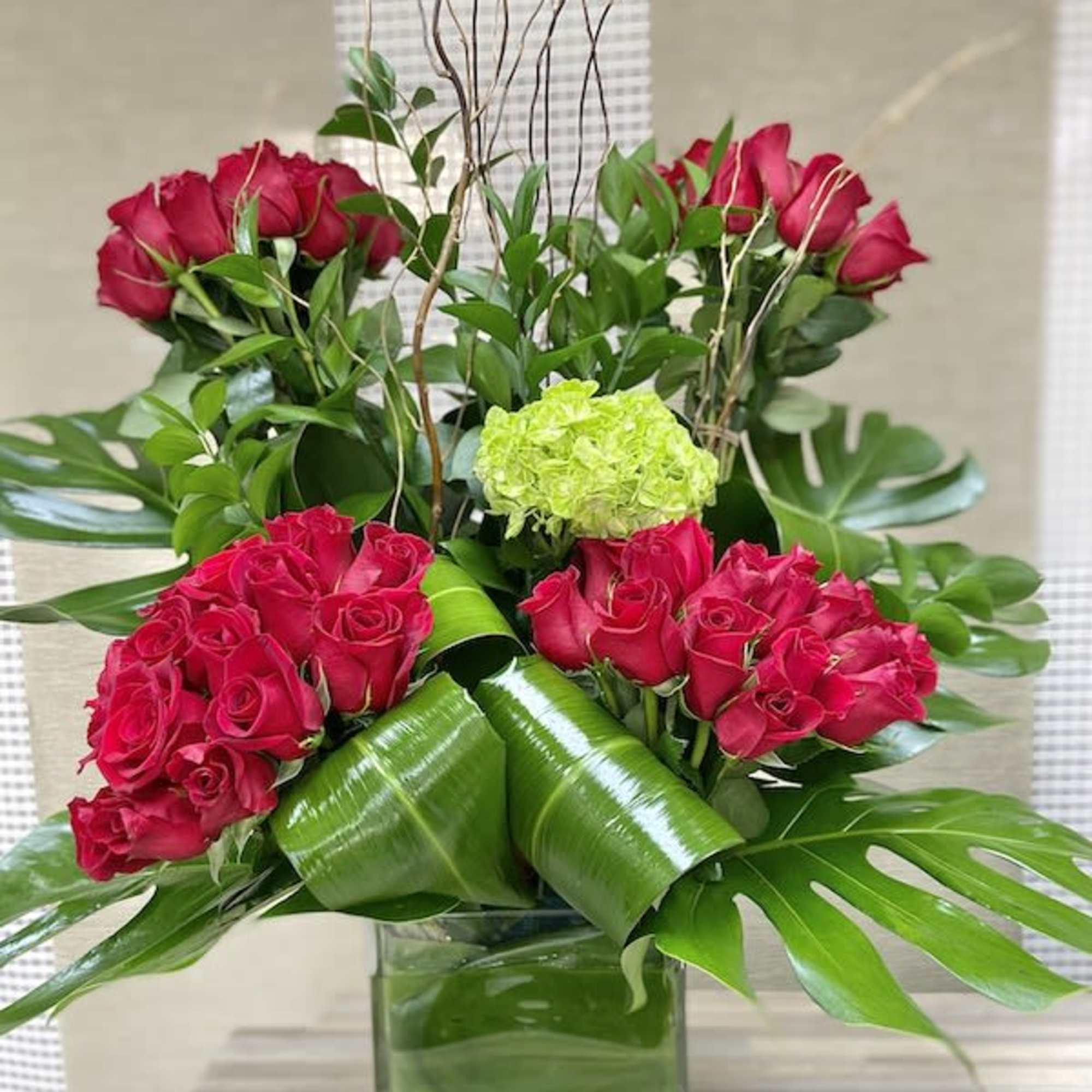 Tall arrangement of red roses with green hydrangea and bold tropical leaves in a glass cube vase