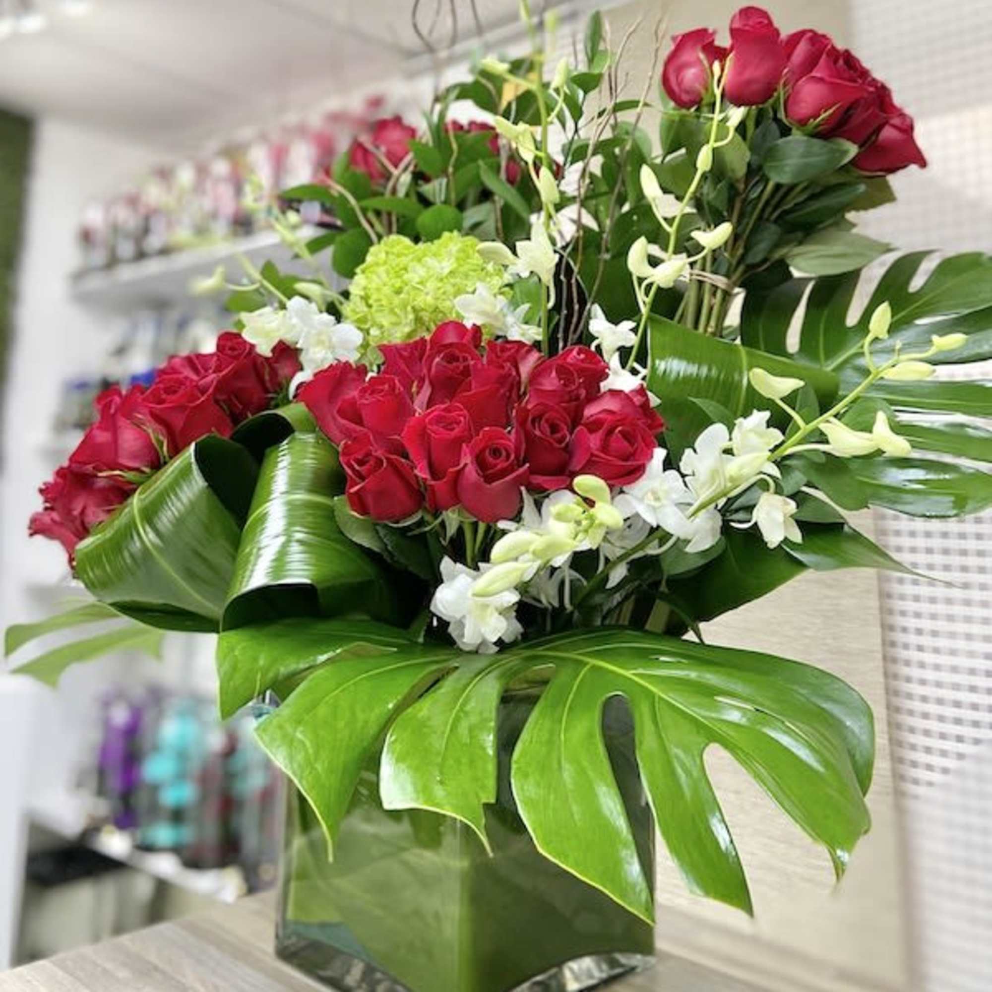 Large arrangement of red roses, white orchids, and green hydrangea in a square glass vase with broad tropical leaves.