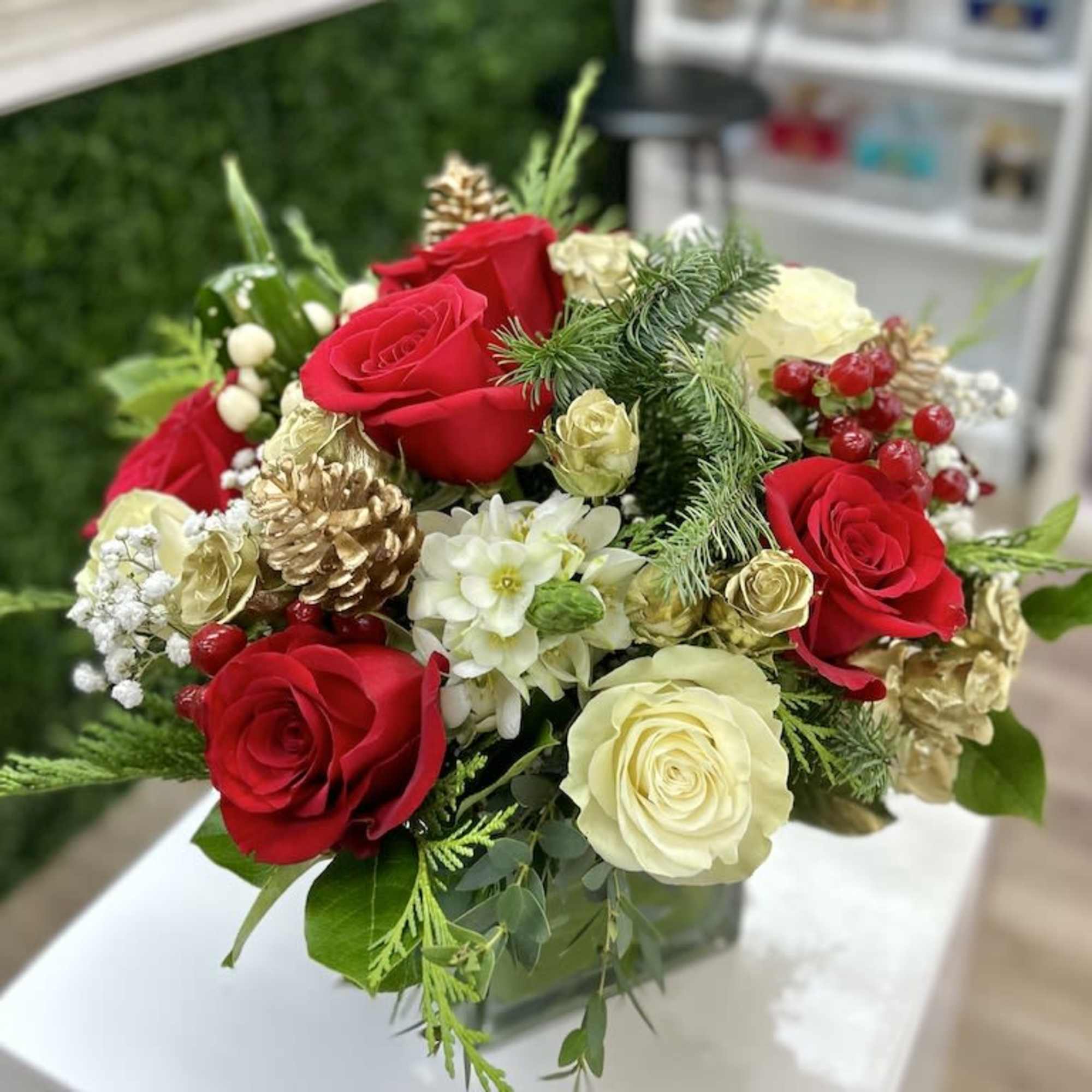Low holiday arrangement with red and cream roses, white blooms, evergreens, and gold pinecones in a glass cube.