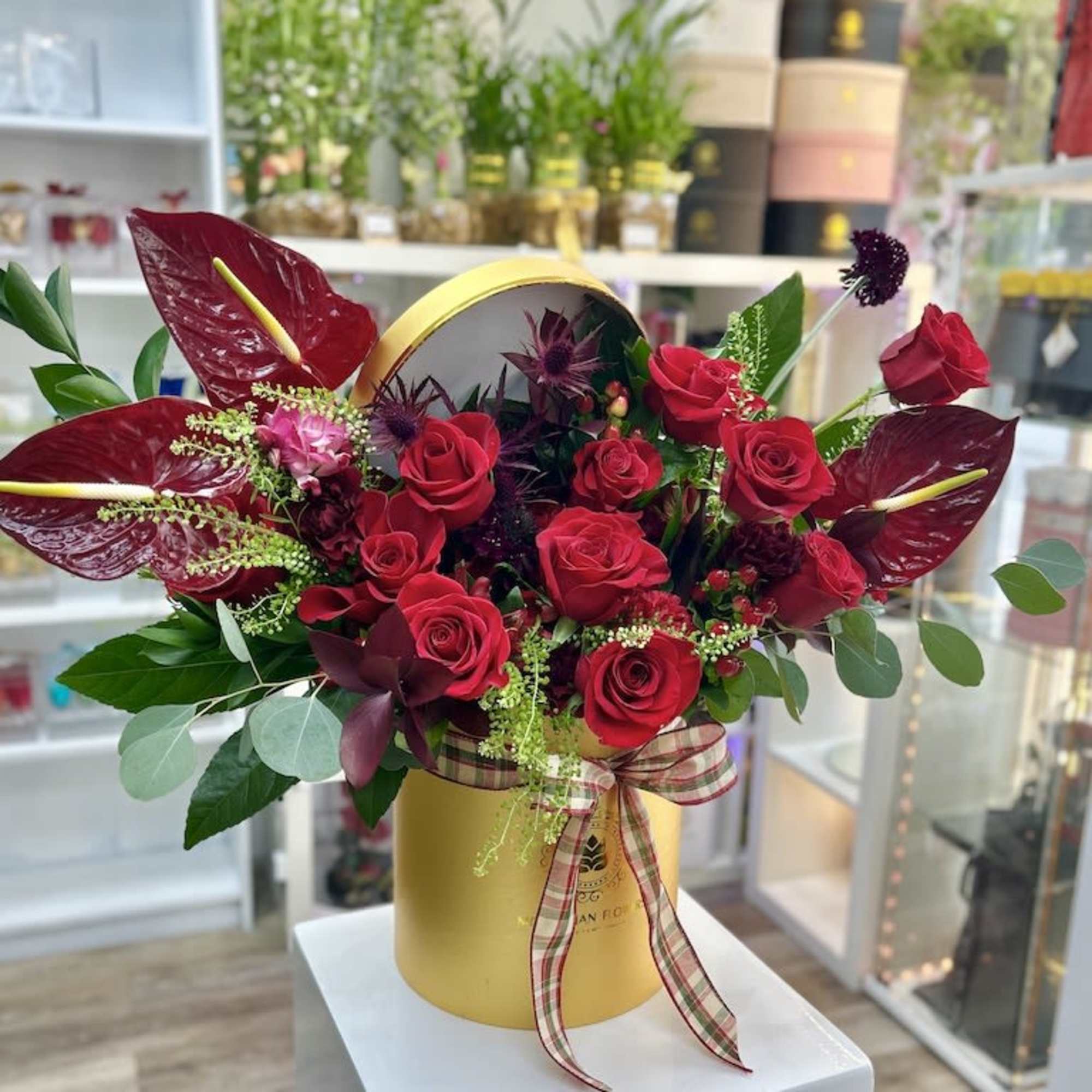 Red rose and tropical flower arrangement in a gold hat box with plaid ribbon.