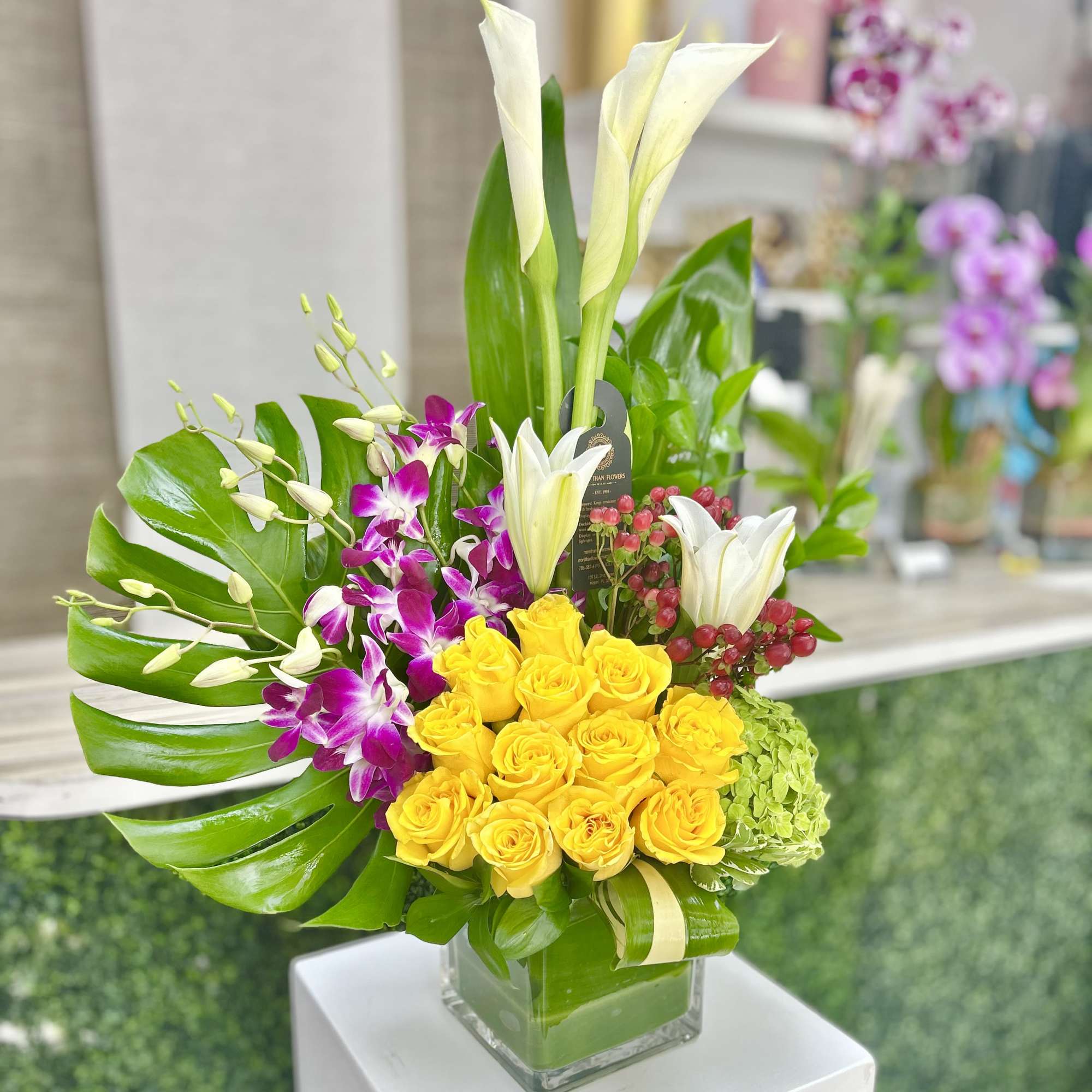 Tall arrangement with yellow roses, white lilies, calla lilies and orchids in a glass cube vase